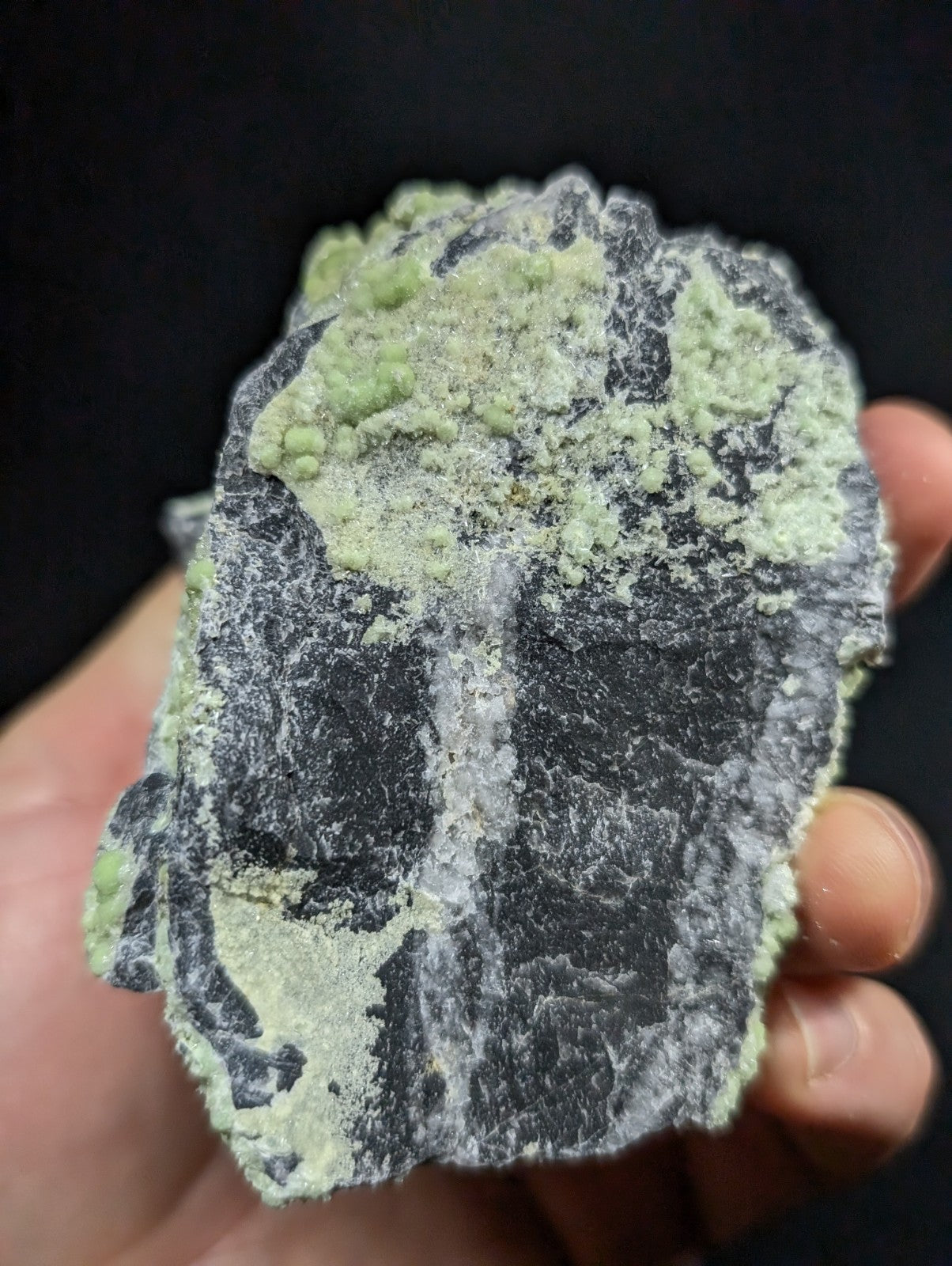 A hand holds a dark gray, rough-textured mineral specimen with patches of light green crystalline growths.