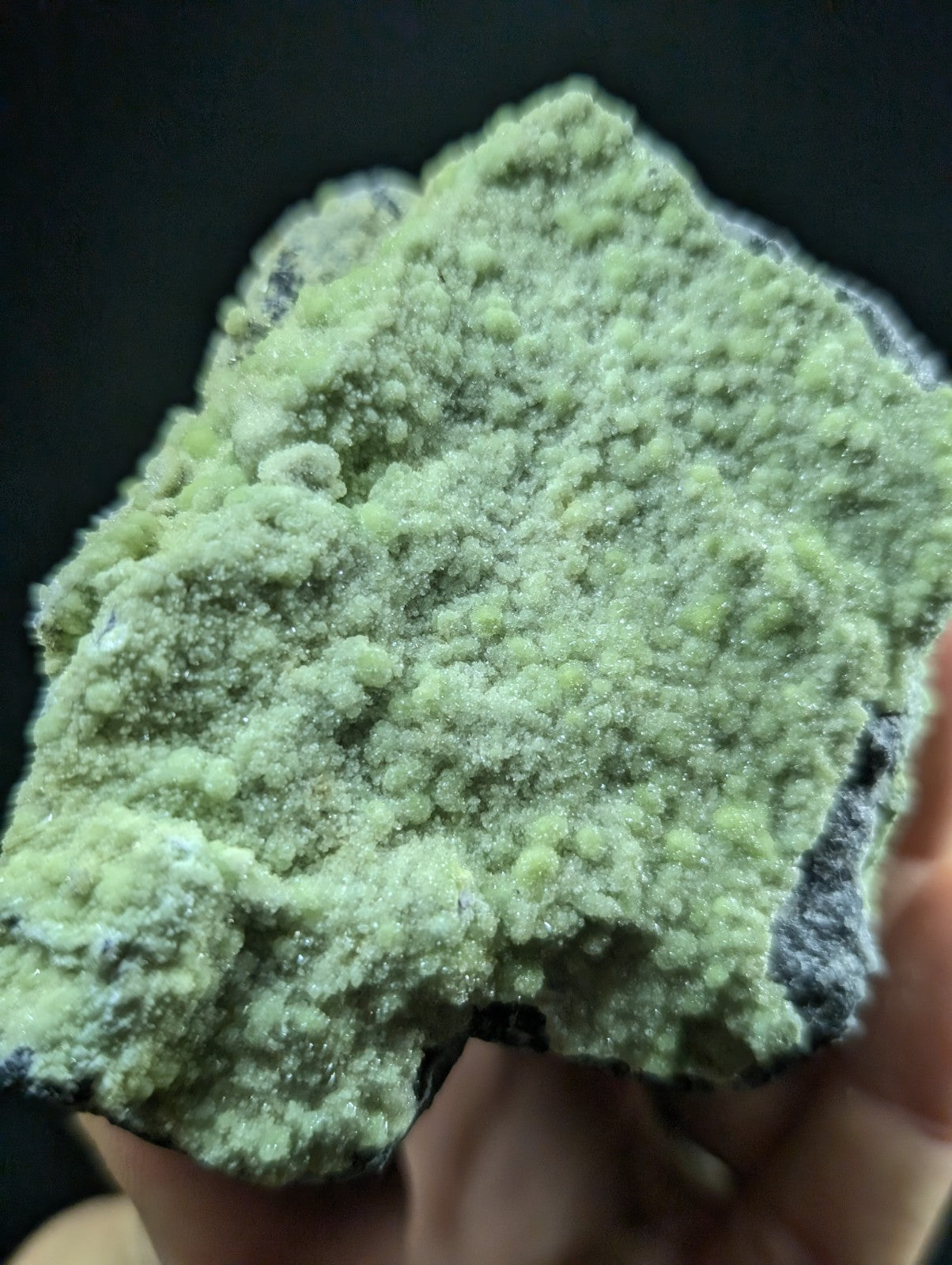 A hand holds a rough, irregularly shaped mineral specimen with vibrant green crystalline formations covering its surface.