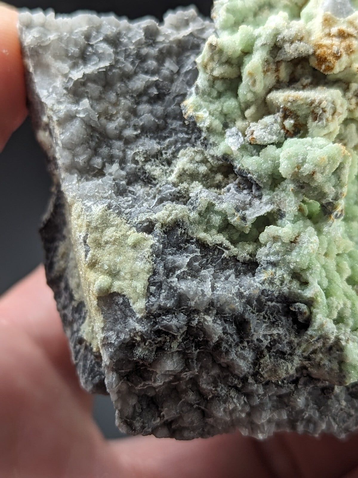 A textured mineral specimen featuring a gray, crystalline base with patches of light green and pale yellowish-green minerals growing in intricate, clustered formations.
