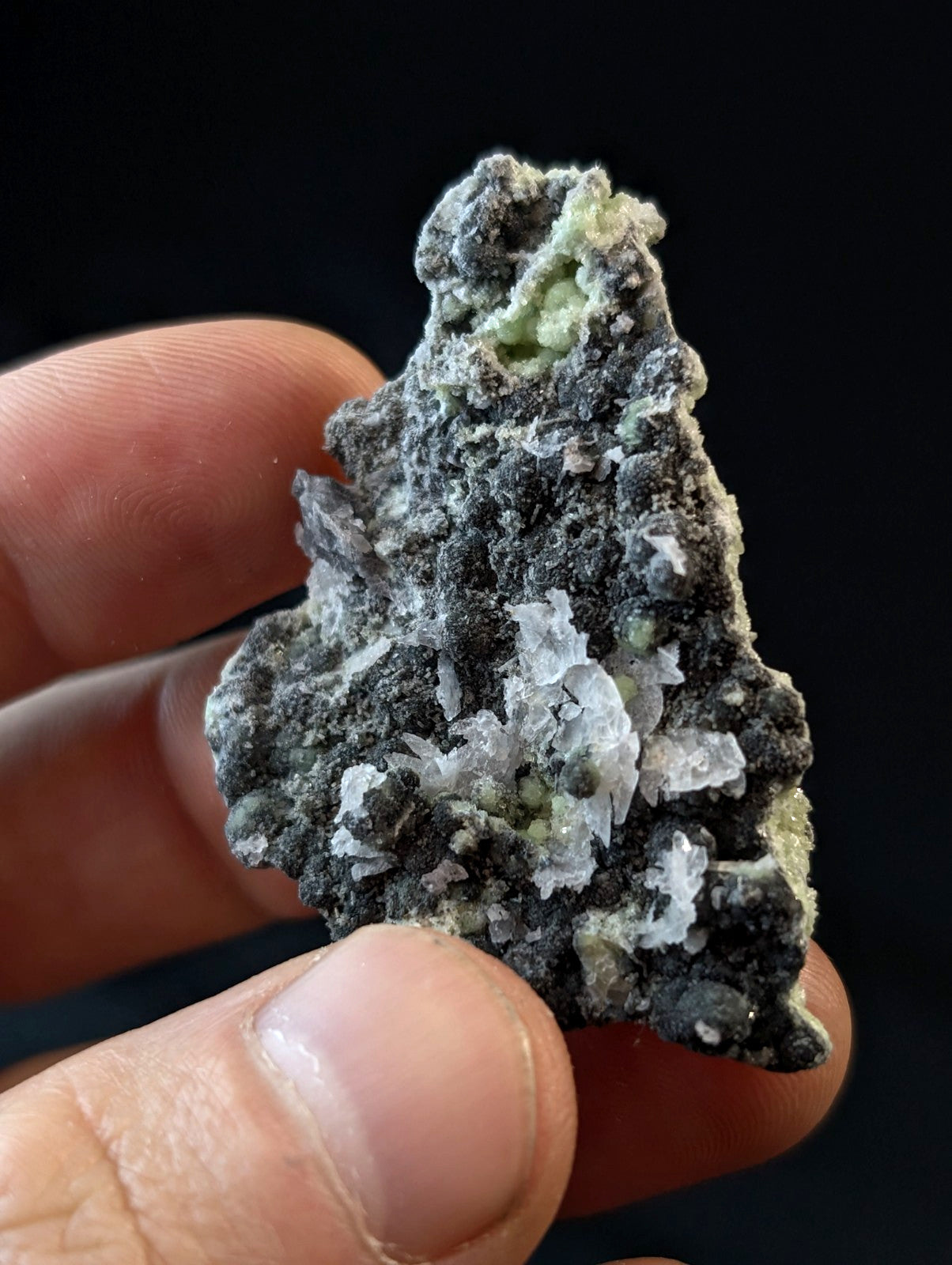 A hand holds a rough, irregularly shaped mineral specimen featuring dark gray matrix with patches of pale green and translucent white crystalline formations.