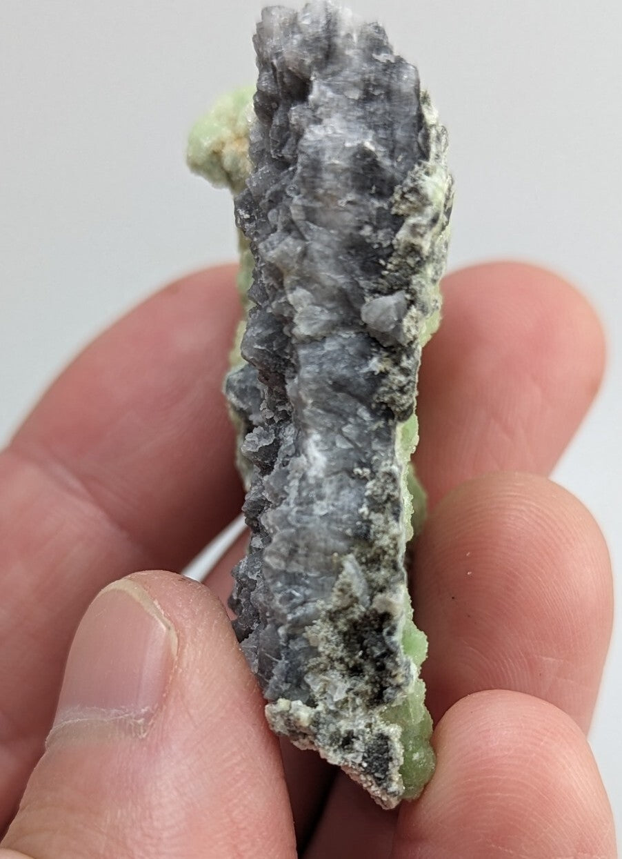 A hand holds a natural mineral specimen featuring dark gray, crystalline formations with patches of light green matrix.