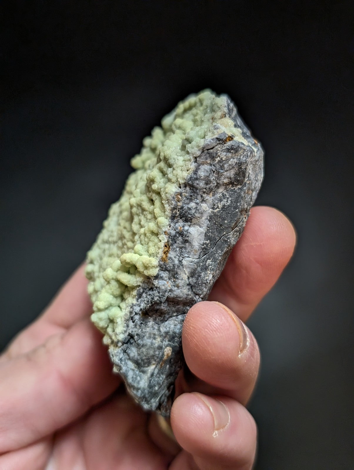 A hand holds a rough, irregularly shaped mineral specimen featuring a striking contrast between its dark gray matrix and vibrant light green crystalline formations.