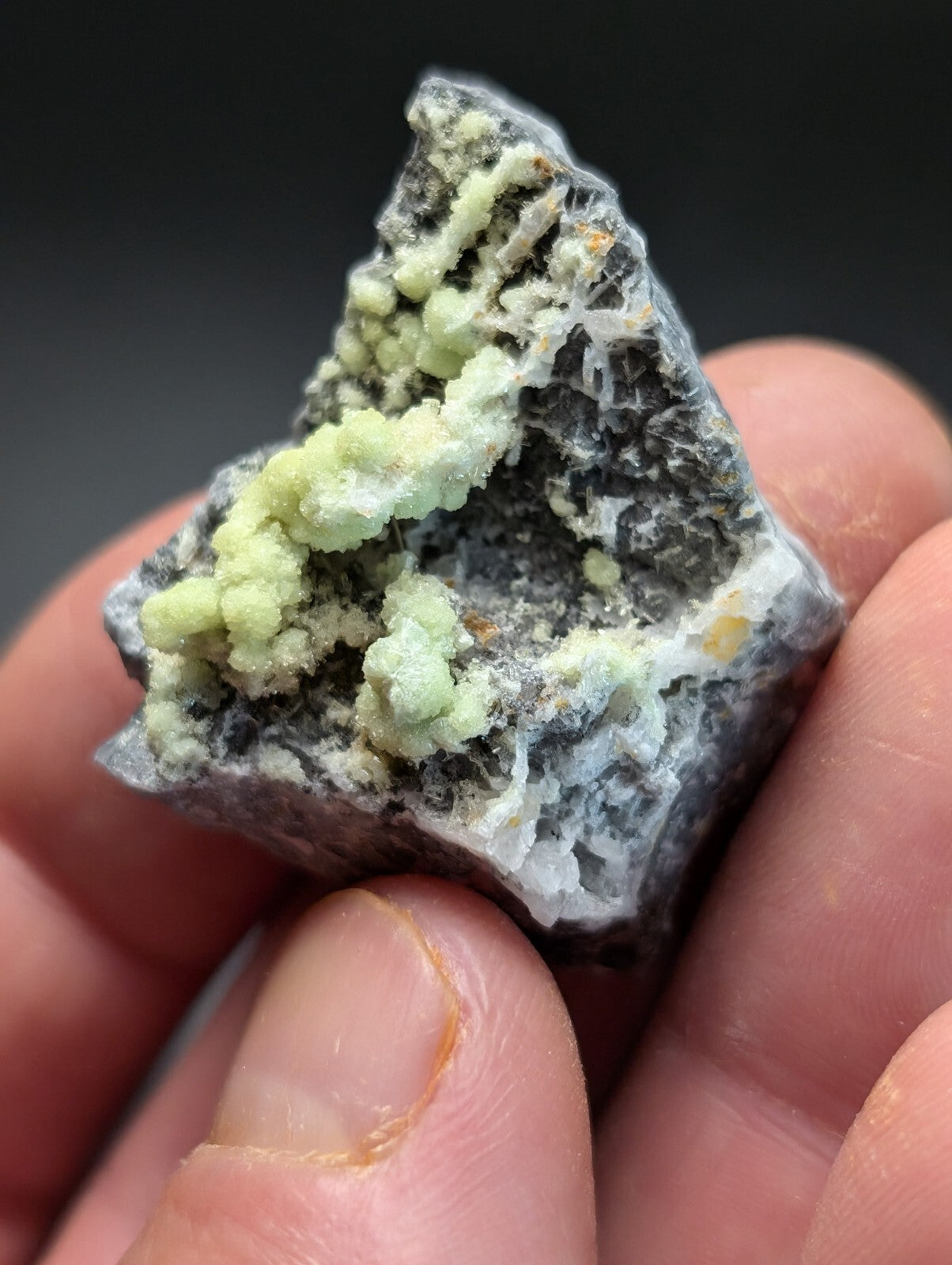 A hand holds a dark gray rock specimen featuring light green, crystalline formations.