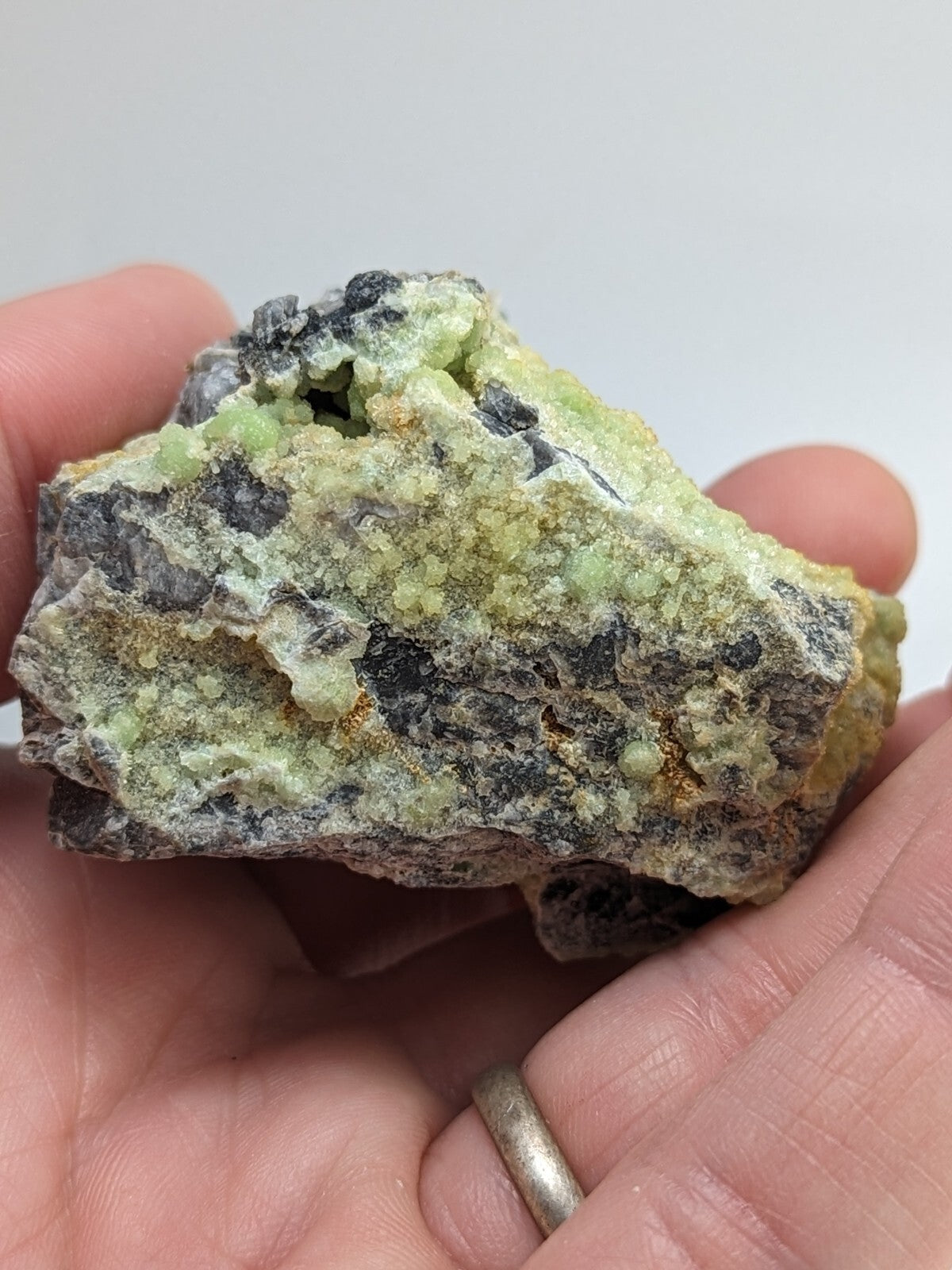 A hand holds a rough, irregularly shaped mineral specimen featuring patches of pale green crystals interspersed with dark gray and black matrix material.