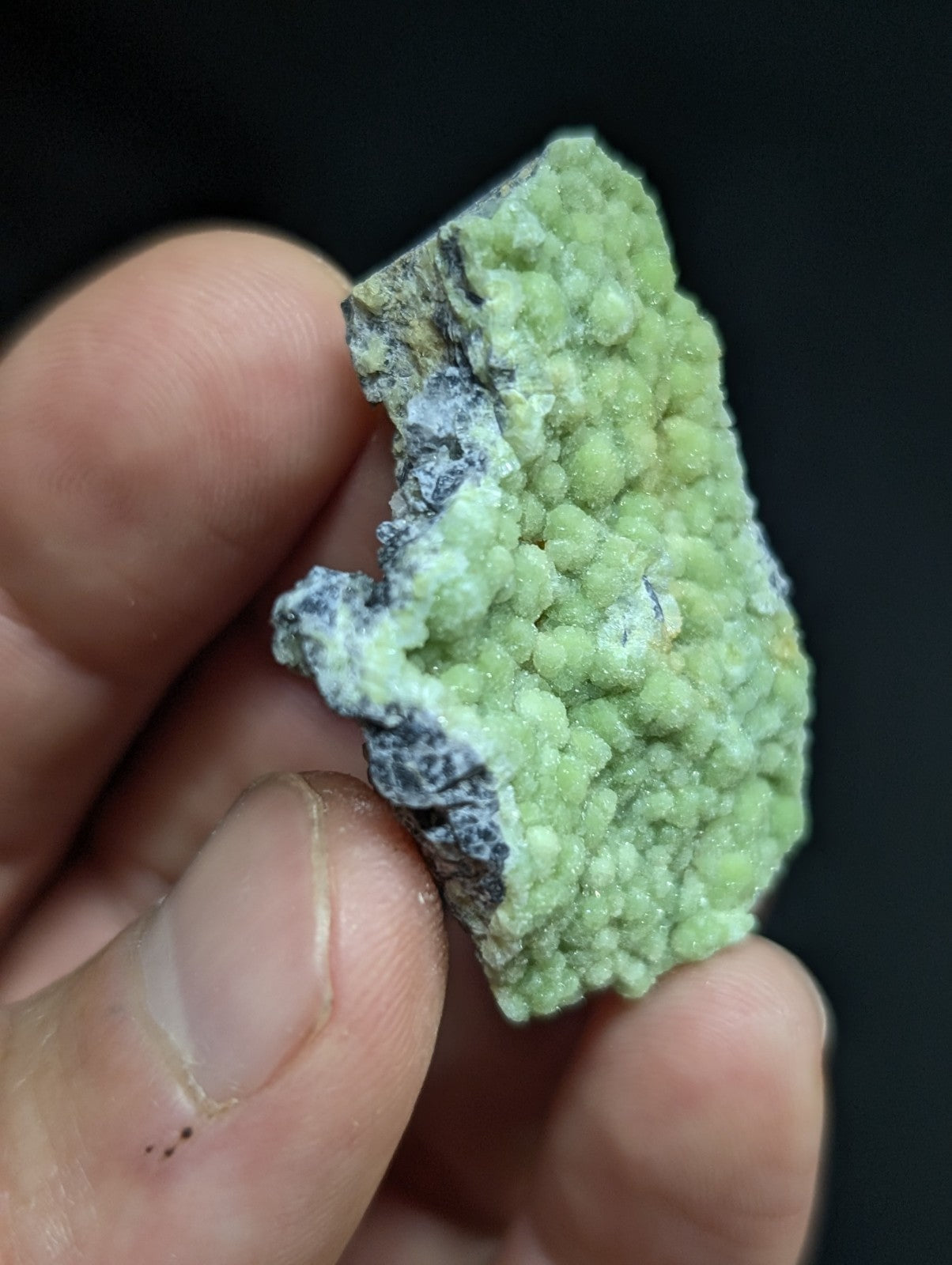 A hand holds a light green, crystalline mineral specimen with rounded, clustered formations and darker grayish matrix.