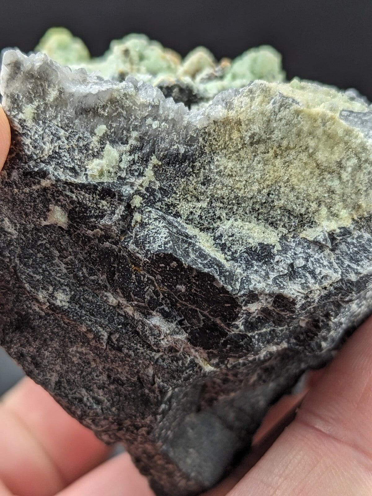 A dark, rough-textured mineral specimen with patches of pale green crystalline growths.