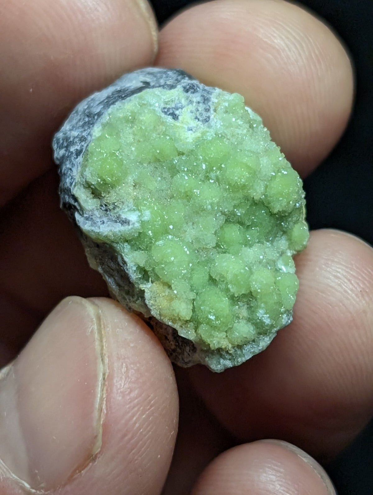 A hand holds a rough, irregularly shaped mineral specimen featuring vibrant green crystalline formations clustered together against a darker gray matrix.