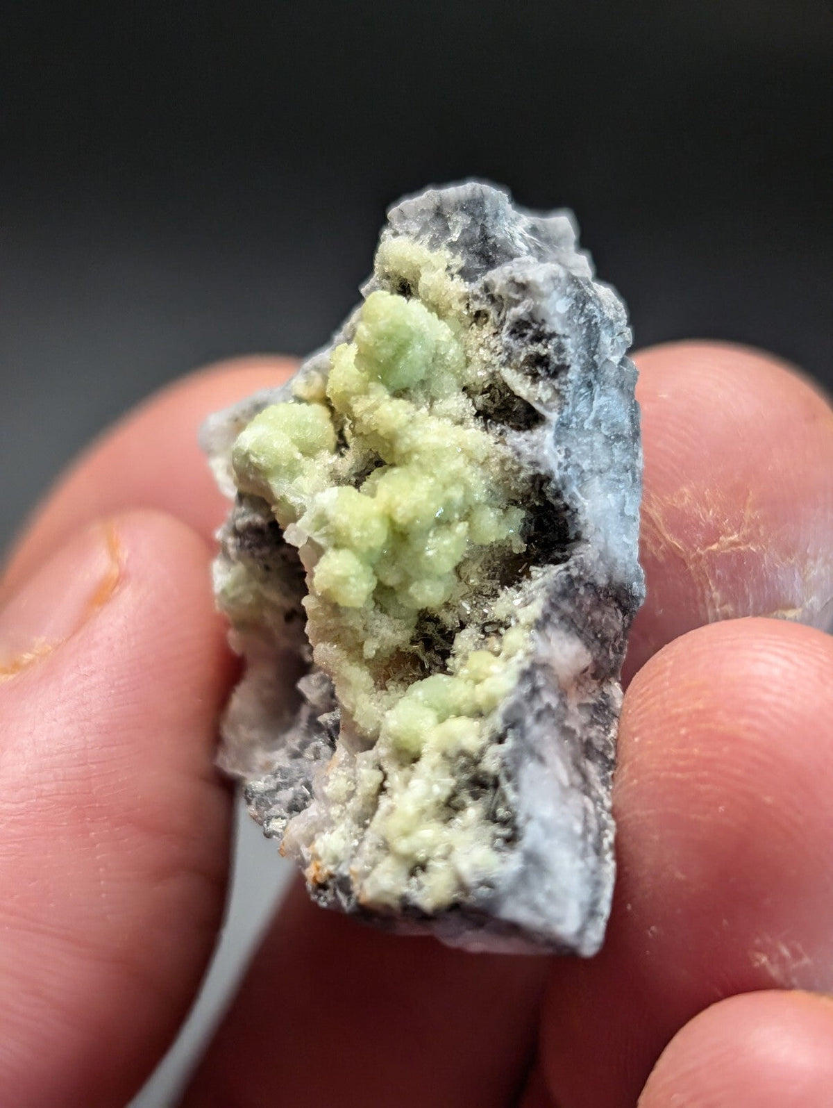 A hand holds a rough, irregularly shaped mineral specimen featuring clusters of pale green crystals growing from a darker gray matrix.