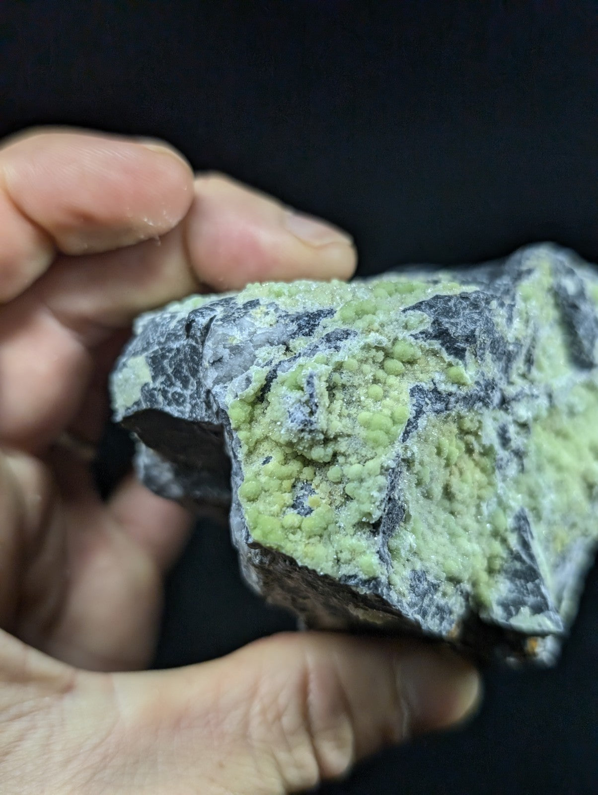 A hand holds a rough, irregularly shaped mineral specimen featuring vibrant green crystalline formations over a dark gray matrix.