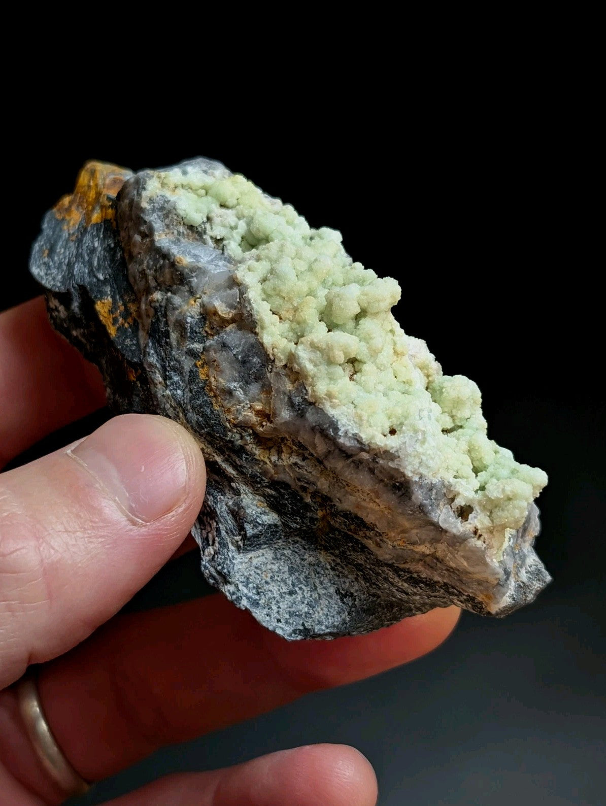 A hand holds a rough, dark gray rock specimen featuring prominent light green crystalline formations.