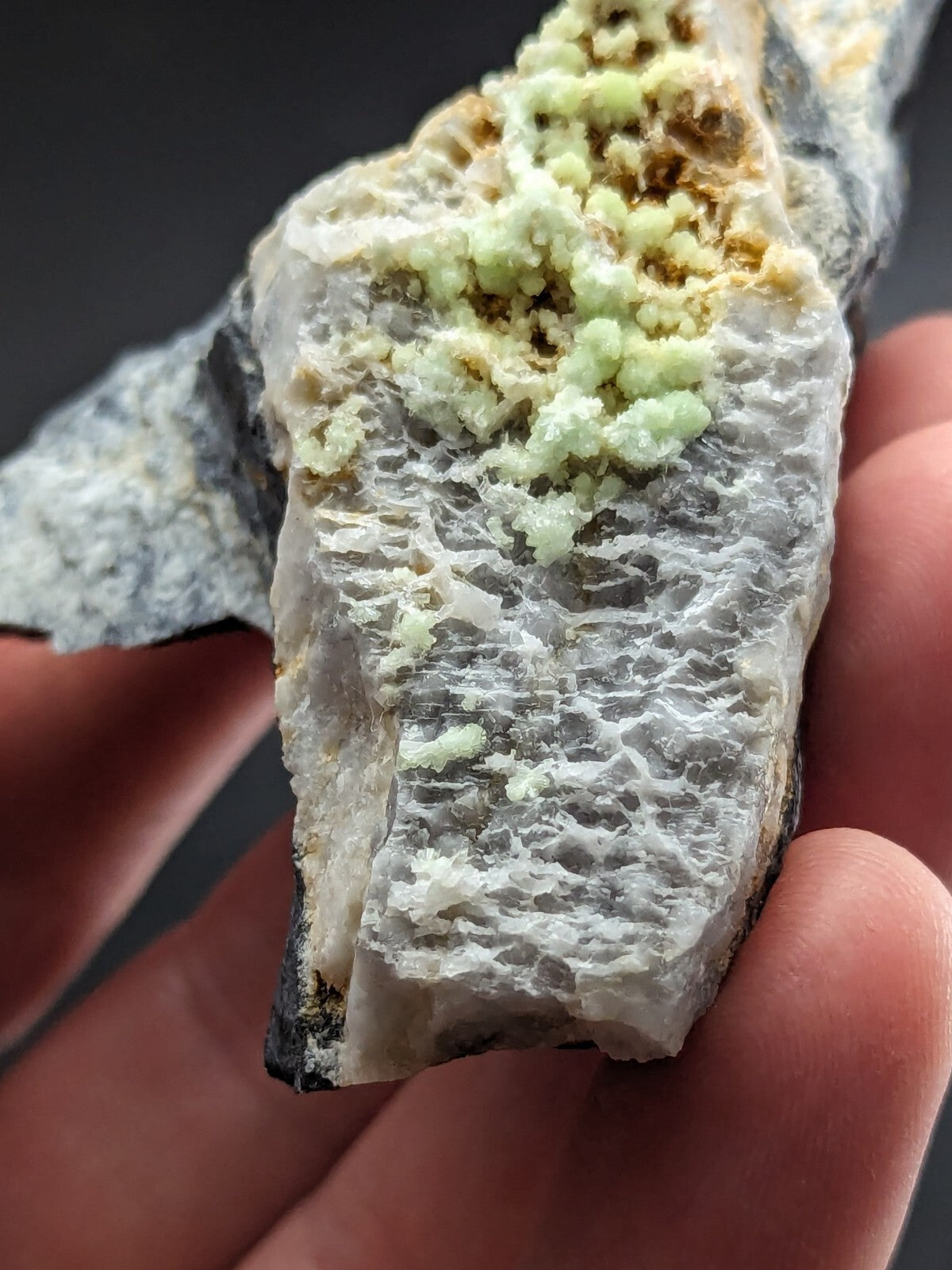 A hand holds a mineral specimen featuring pale green, crystalline formations growing atop a textured white and gray rock matrix.