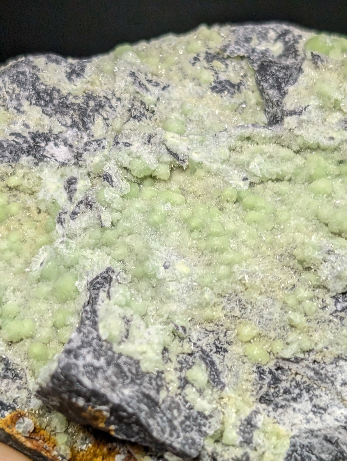 A specimen of greenish-yellow malachite with a crystalline, bead-like texture and patches of dark gray matrix.