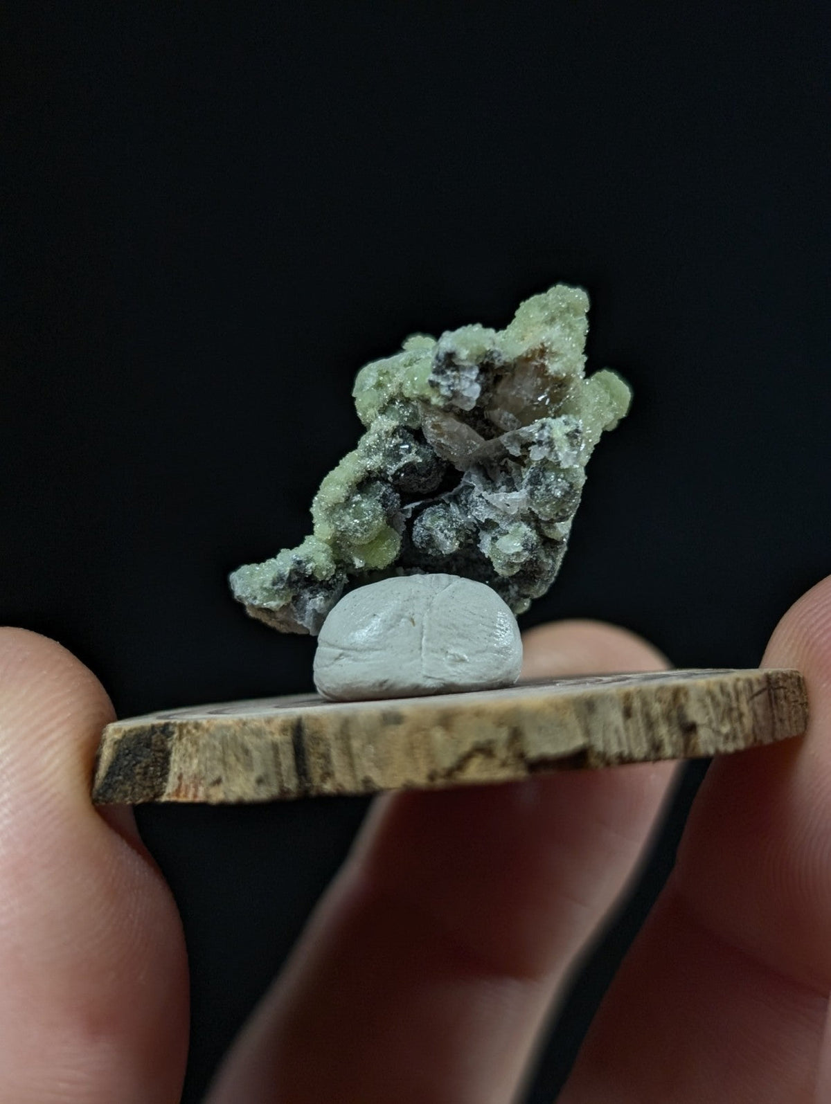 A green and gray mineral specimen with a rough, crystalline texture rests atop a smooth white stone on a rustic wooden slice.