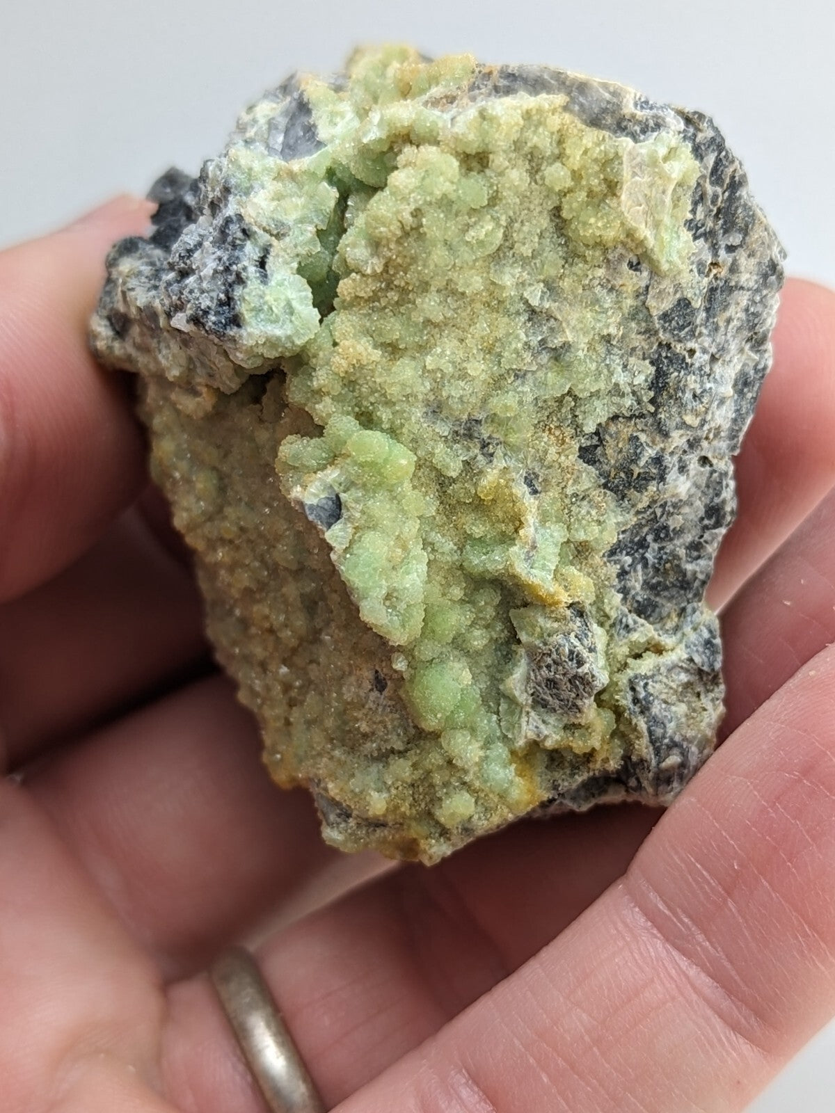 A hand holds a rough, irregularly shaped mineral specimen featuring vibrant green crystals growing from a darker gray matrix.