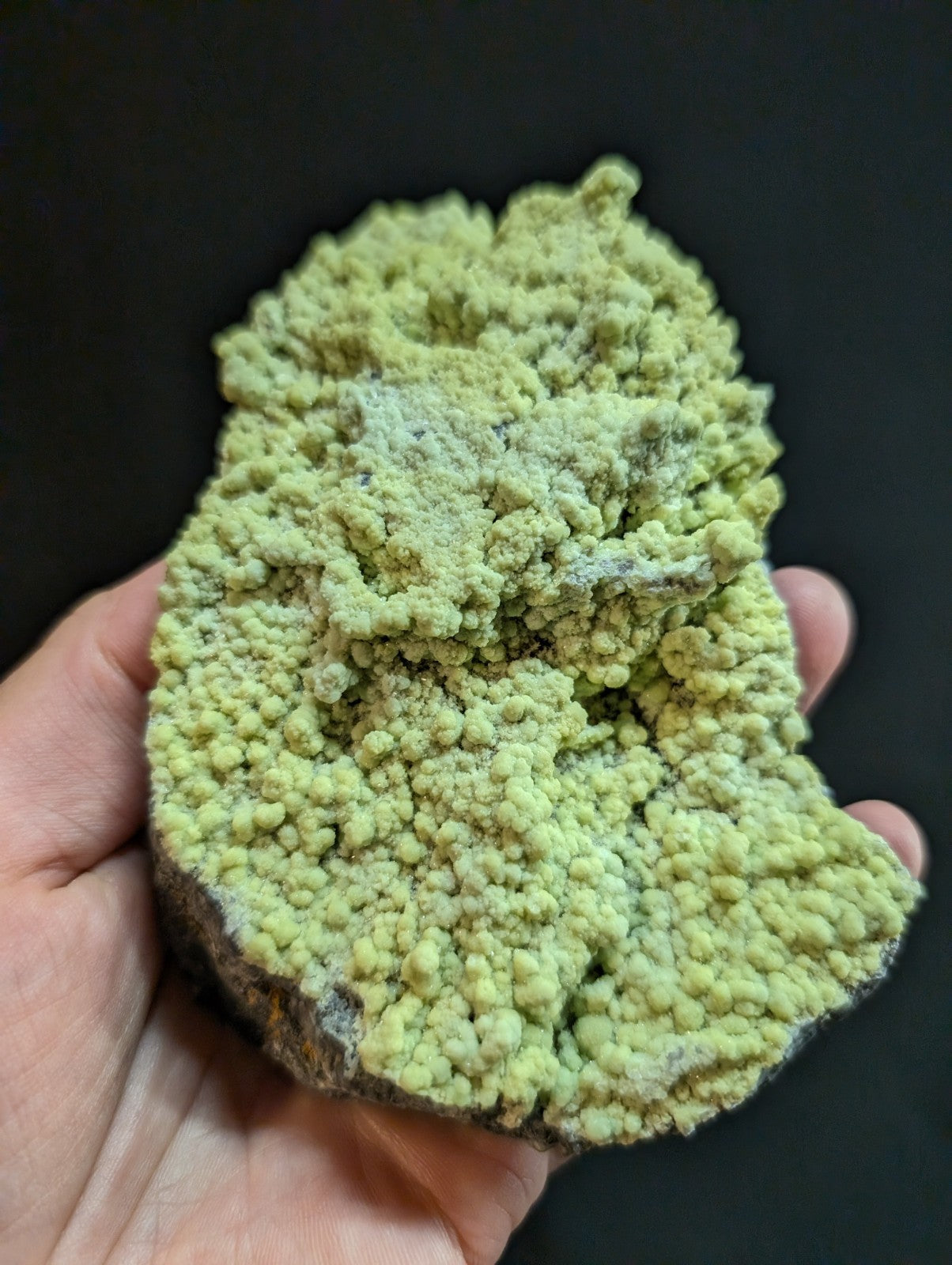 A hand holds a rough, irregularly shaped mineral specimen with vibrant lime-green crystalline formations covering its surface.