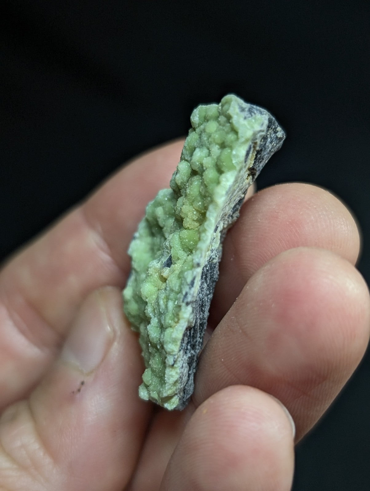 A hand holds a small, irregularly shaped mineral specimen featuring vibrant green crystalline formations clustered along its surface against a dark base.