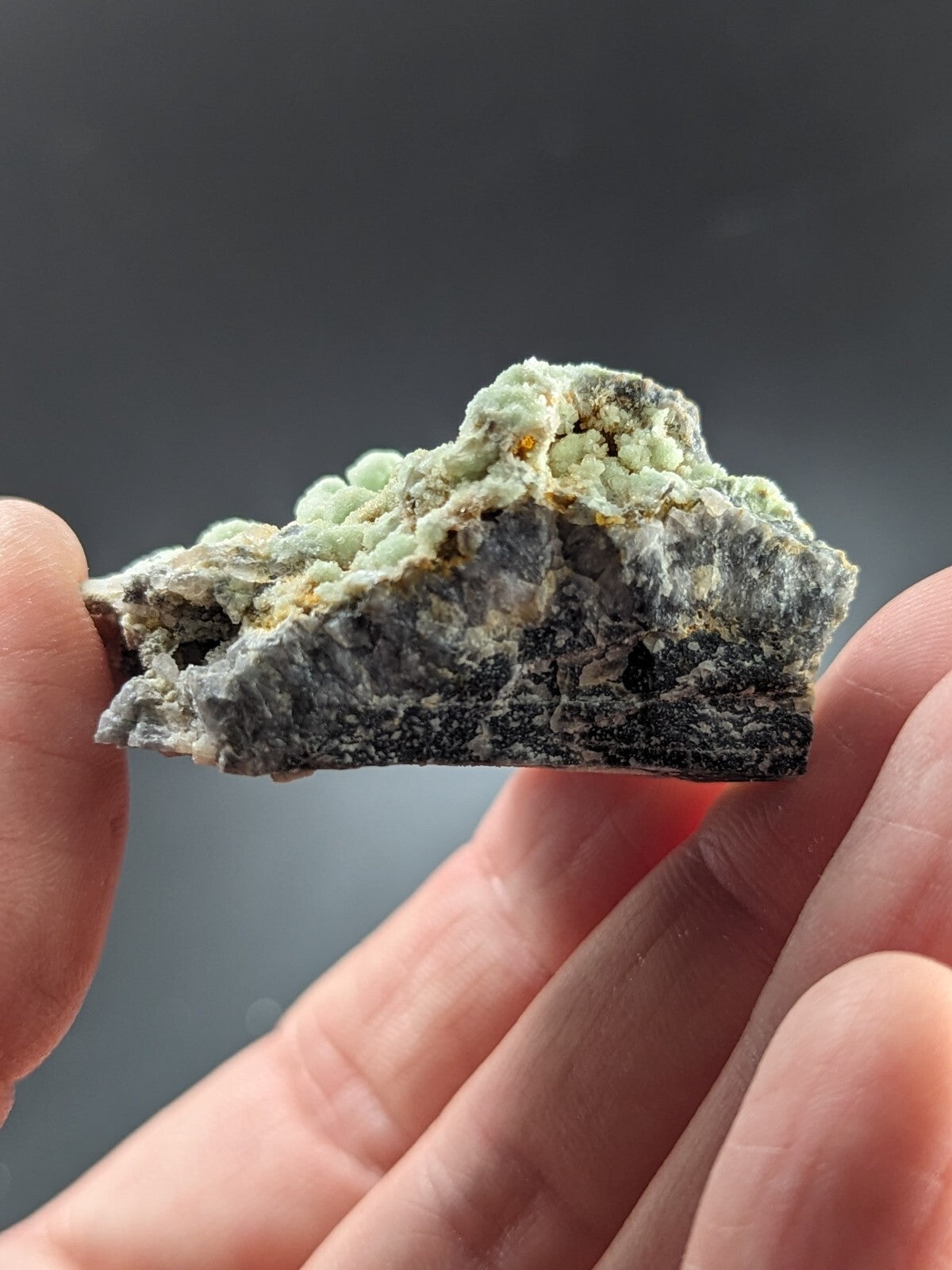 A hand holds a rough, irregularly shaped mineral specimen featuring light green crystalline formations atop a darker gray matrix with subtle yellowish accents.