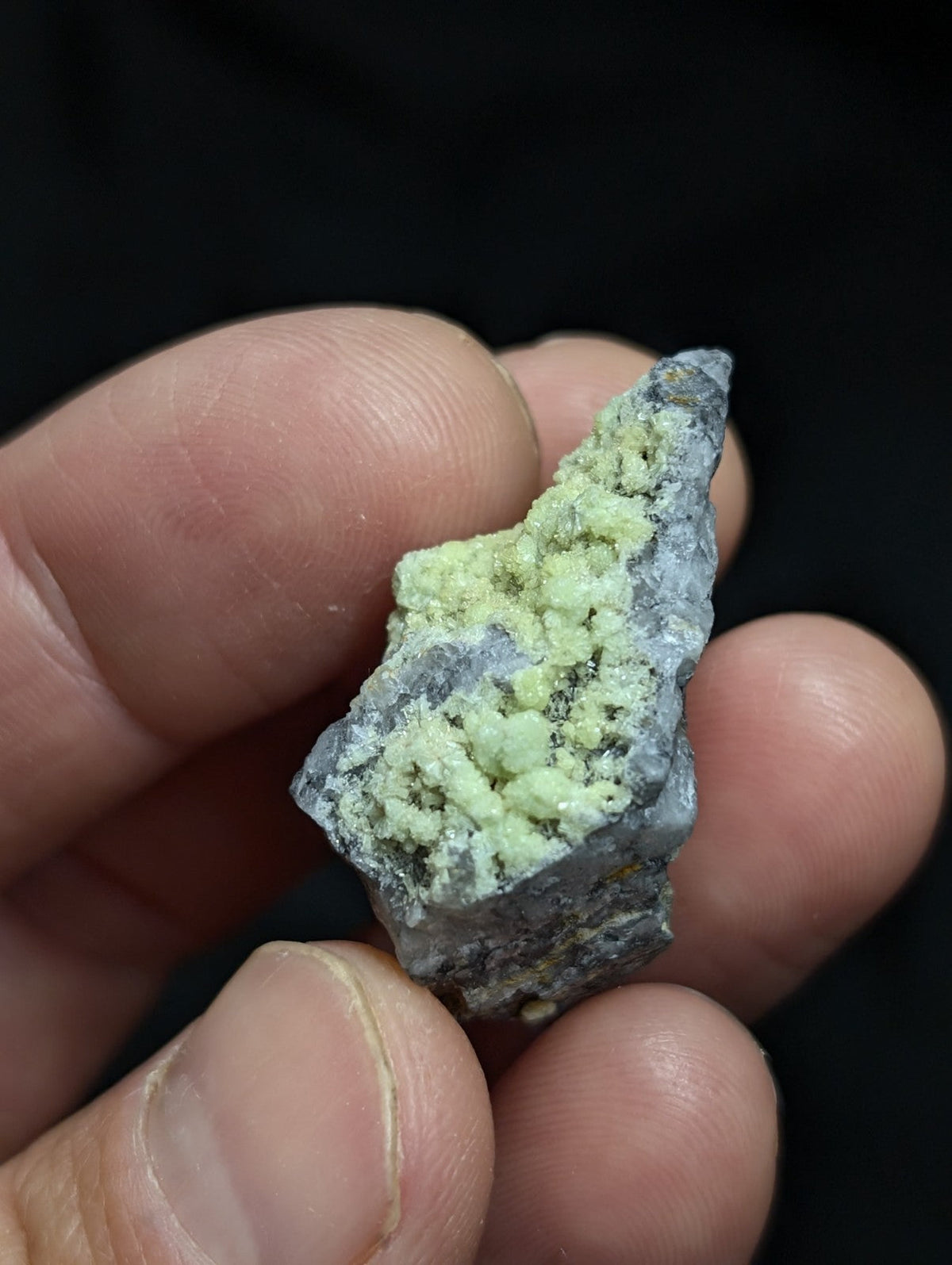 A hand holds a rough, irregularly shaped mineral specimen featuring a grayish base with prominent light green crystalline growths.