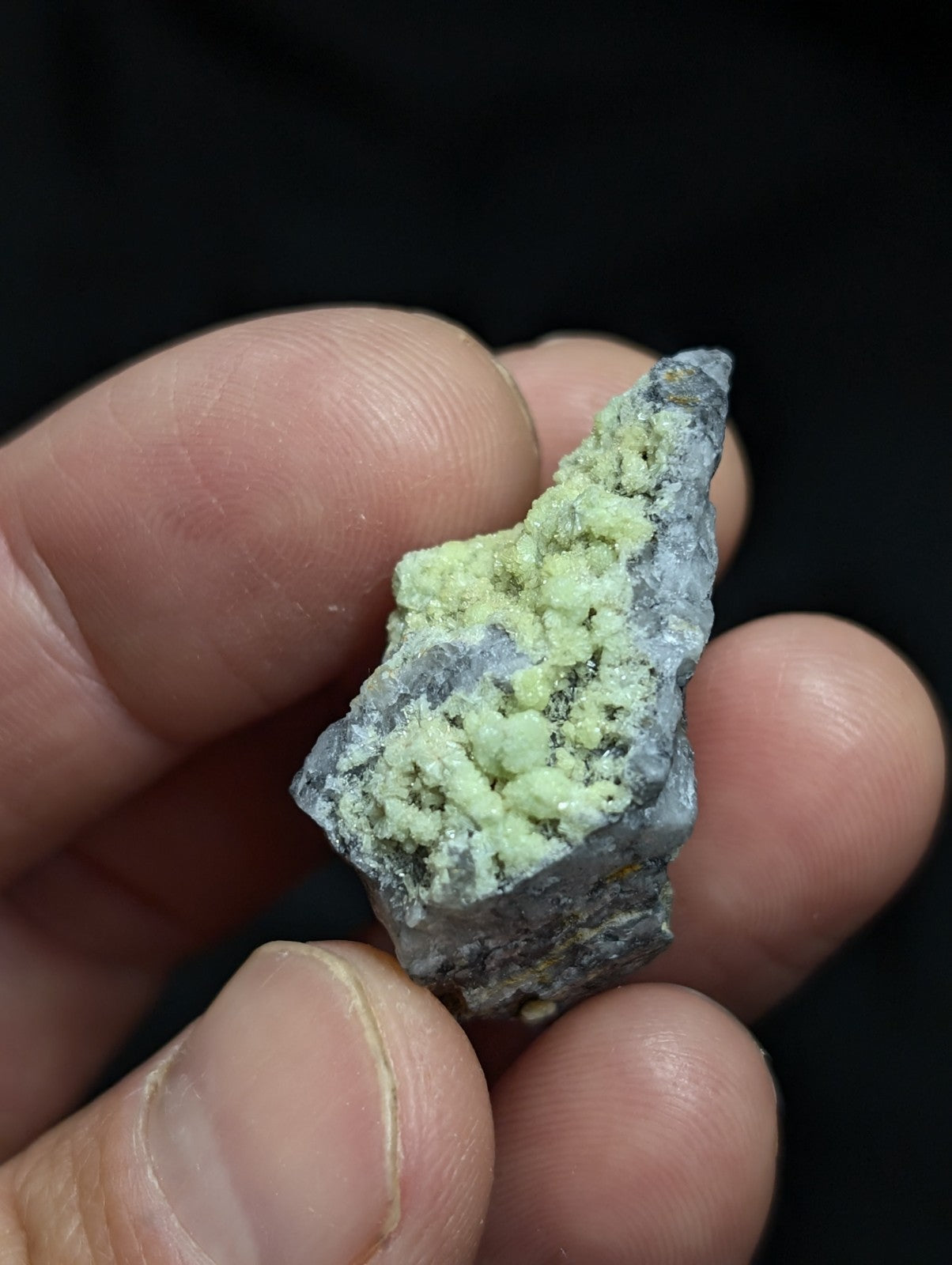 A hand holds a rough, irregularly shaped mineral specimen featuring a grayish base with prominent light green crystalline growths.