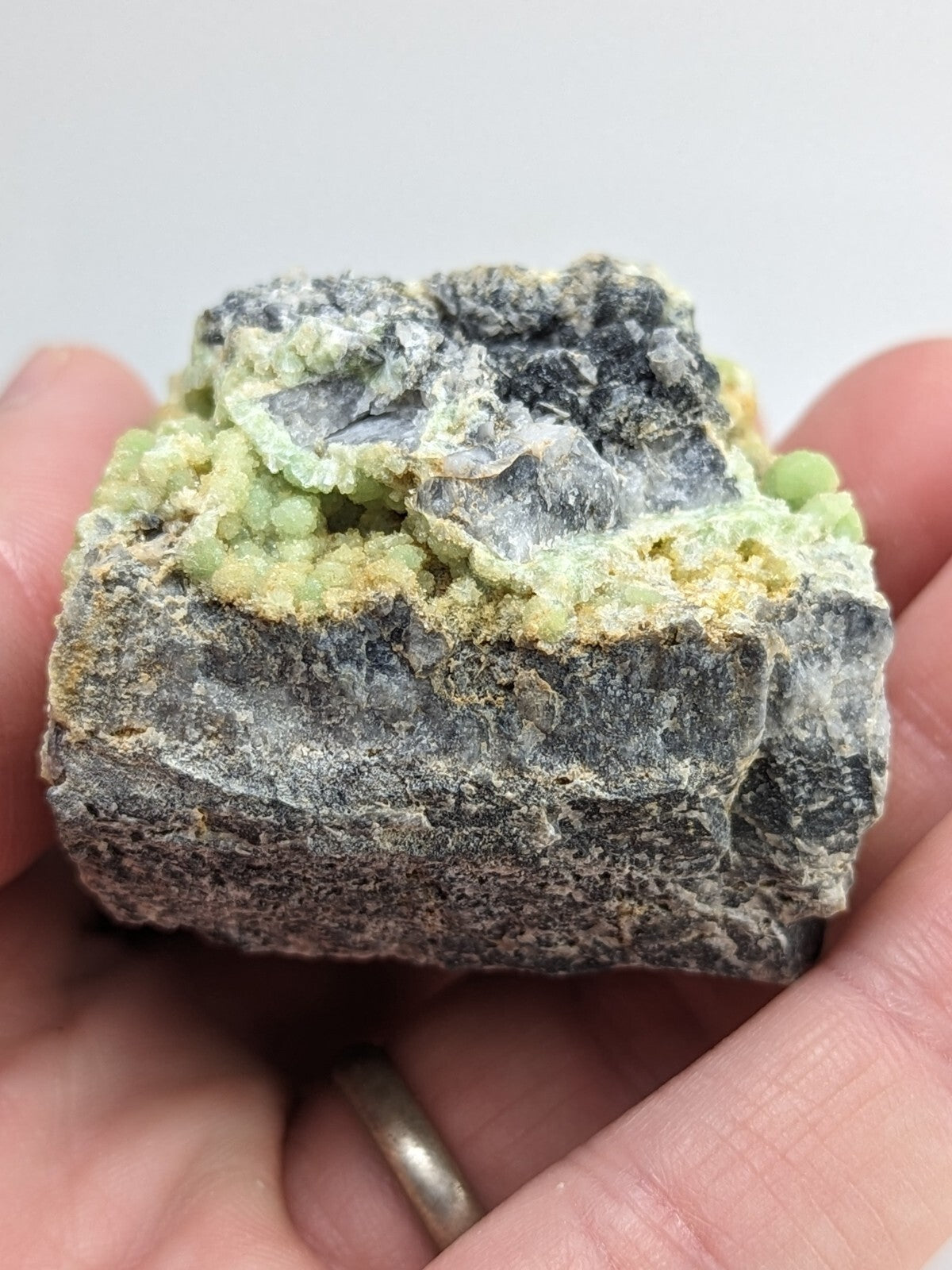 A hand holds a rough, dark gray rock specimen featuring clusters of vibrant light green crystals growing from its surface.