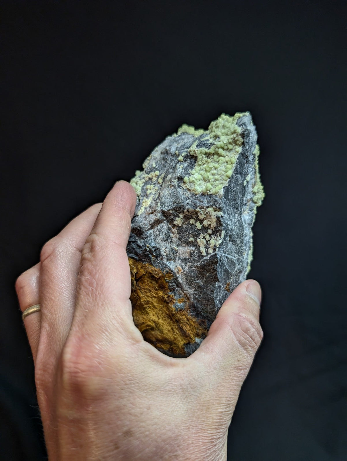 A hand holds a rugged, naturally occurring mineral specimen featuring patches of pale green and golden-yellow crystalline formations against a dark gray matrix.