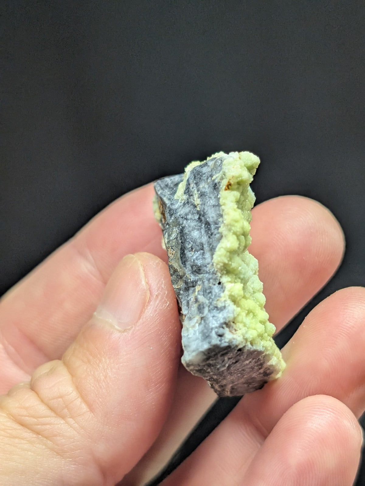 A hand holds a small, irregularly shaped mineral specimen featuring a dark gray base with vibrant green crystalline growths along its edge.
