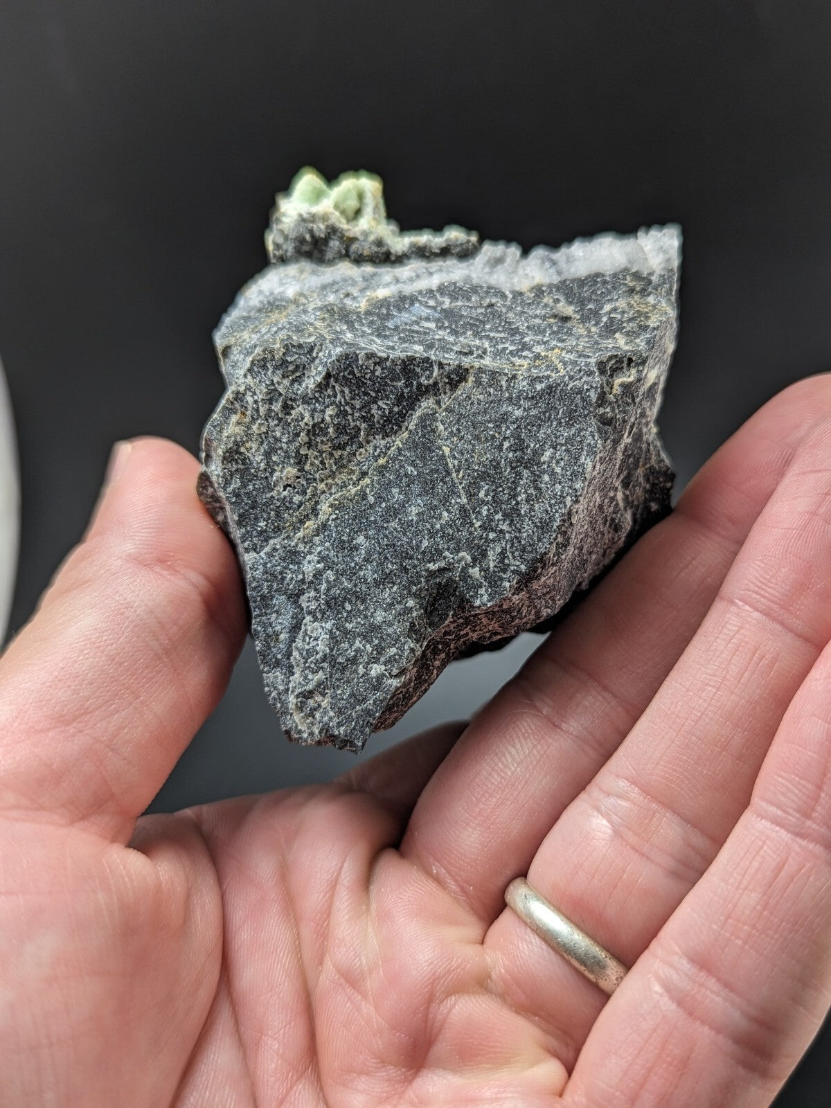 A dark gray, rough-textured rock specimen with a small cluster of light green mineral growths on its upper surface.