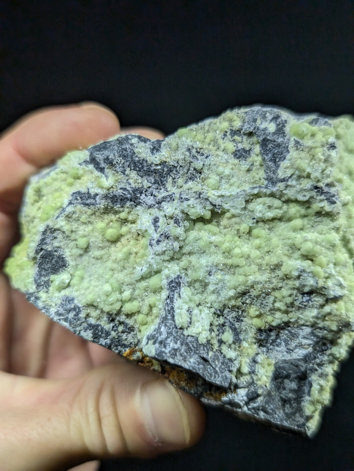 A hand holds a rough, irregularly shaped mineral specimen featuring vibrant lime green and dark gray patches with a textured surface.