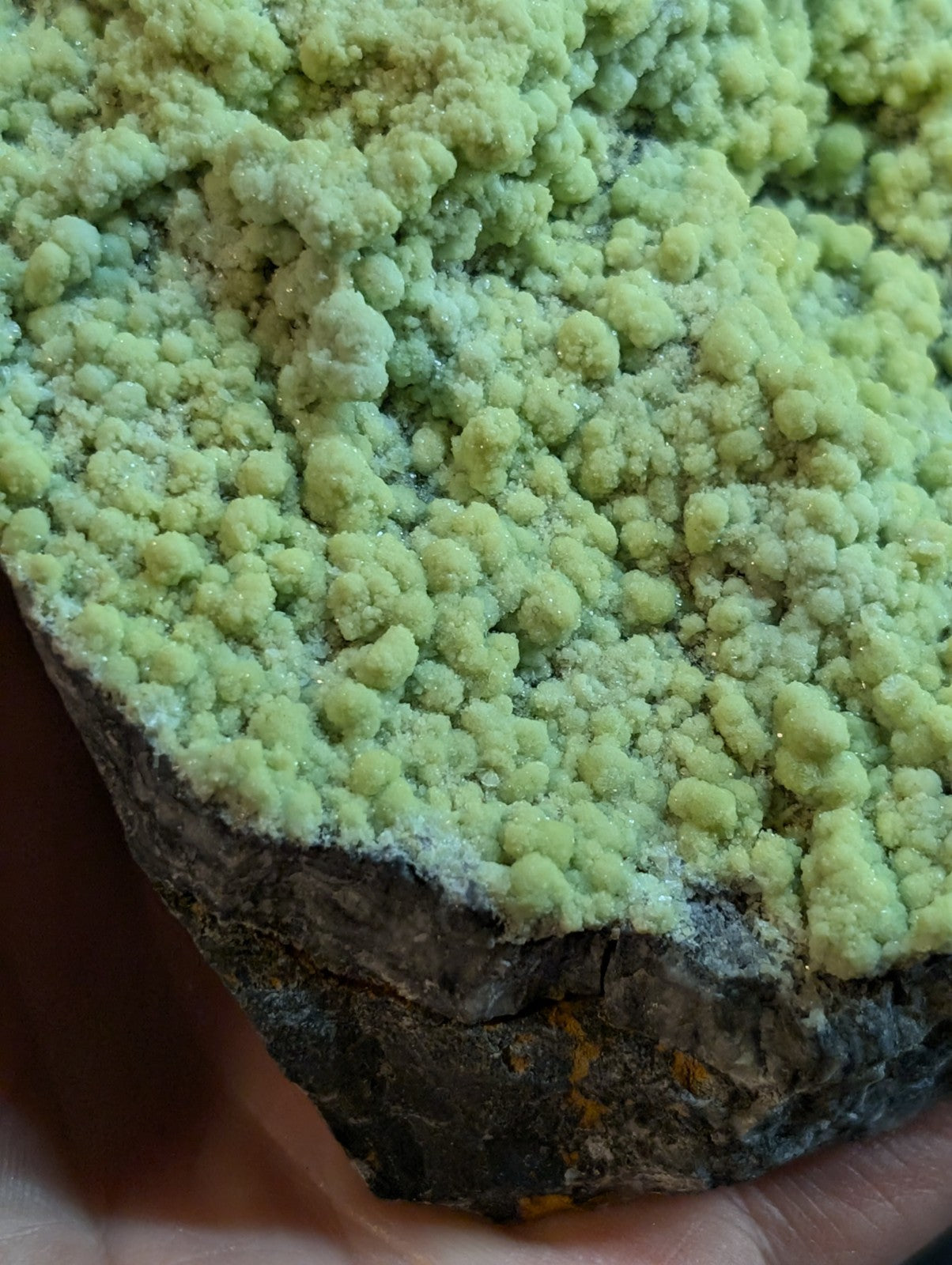 A vibrant lime-green, crystalline mineral formation with a rough, textured surface growing from a dark, rocky base.
