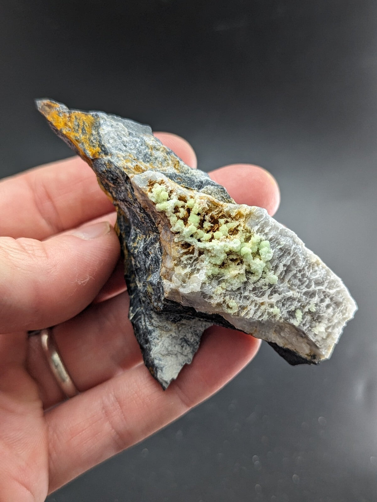 A hand holds a striking mineral specimen featuring a dark, textured base with vibrant green crystalline formations and hints of yellow at its tip.