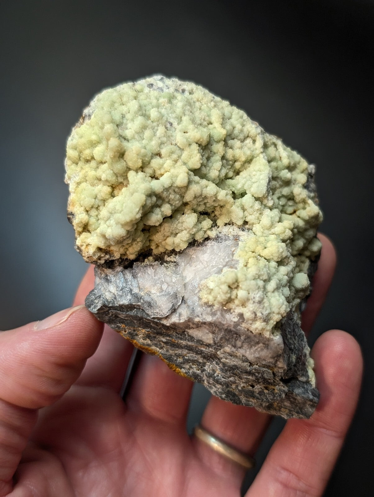 A hand holds a rough, irregularly shaped mineral specimen featuring dense clusters of pale greenish-yellow crystals growing over a darker gray rock base.
