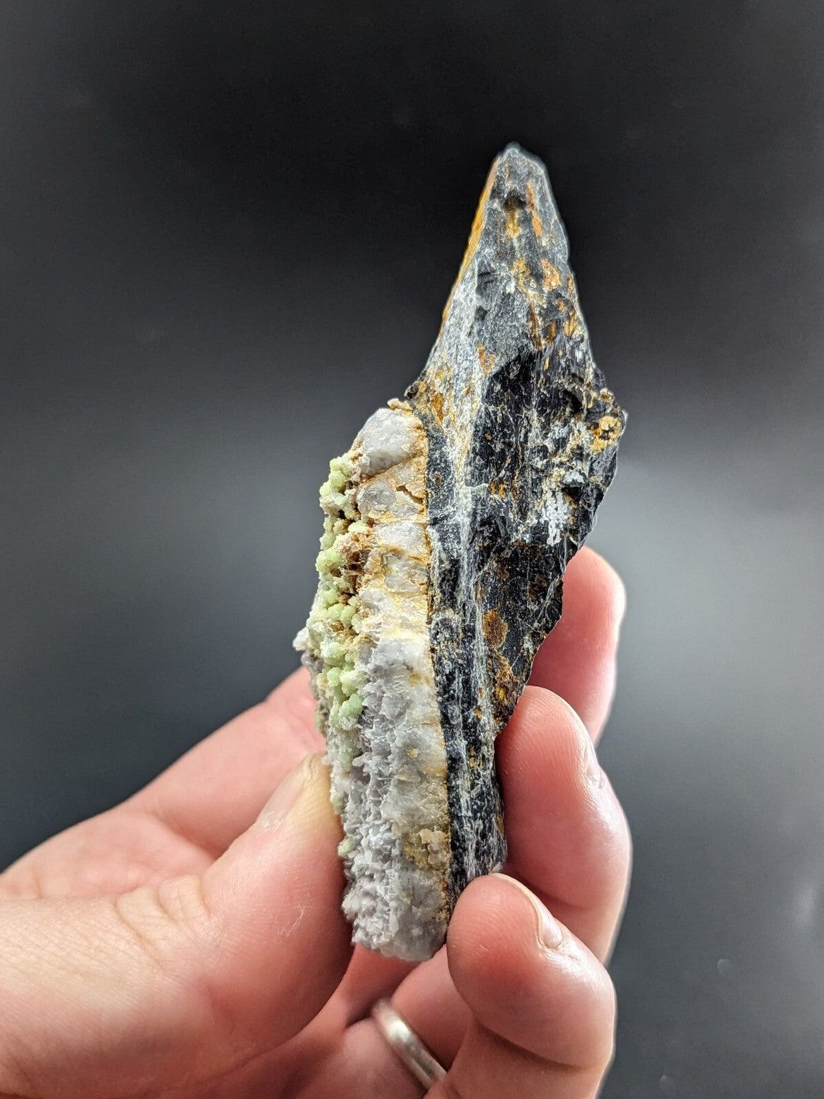 A hand holds a dark, jagged mineral specimen with patches of green and white crystalline growths along its side.