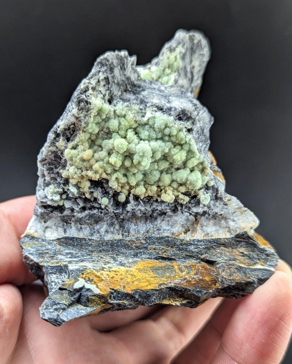 A hand holds a striking geological specimen featuring a cluster of light green, crystalline formations atop a dark gray rock base with streaks of yellow and white mineral veins.