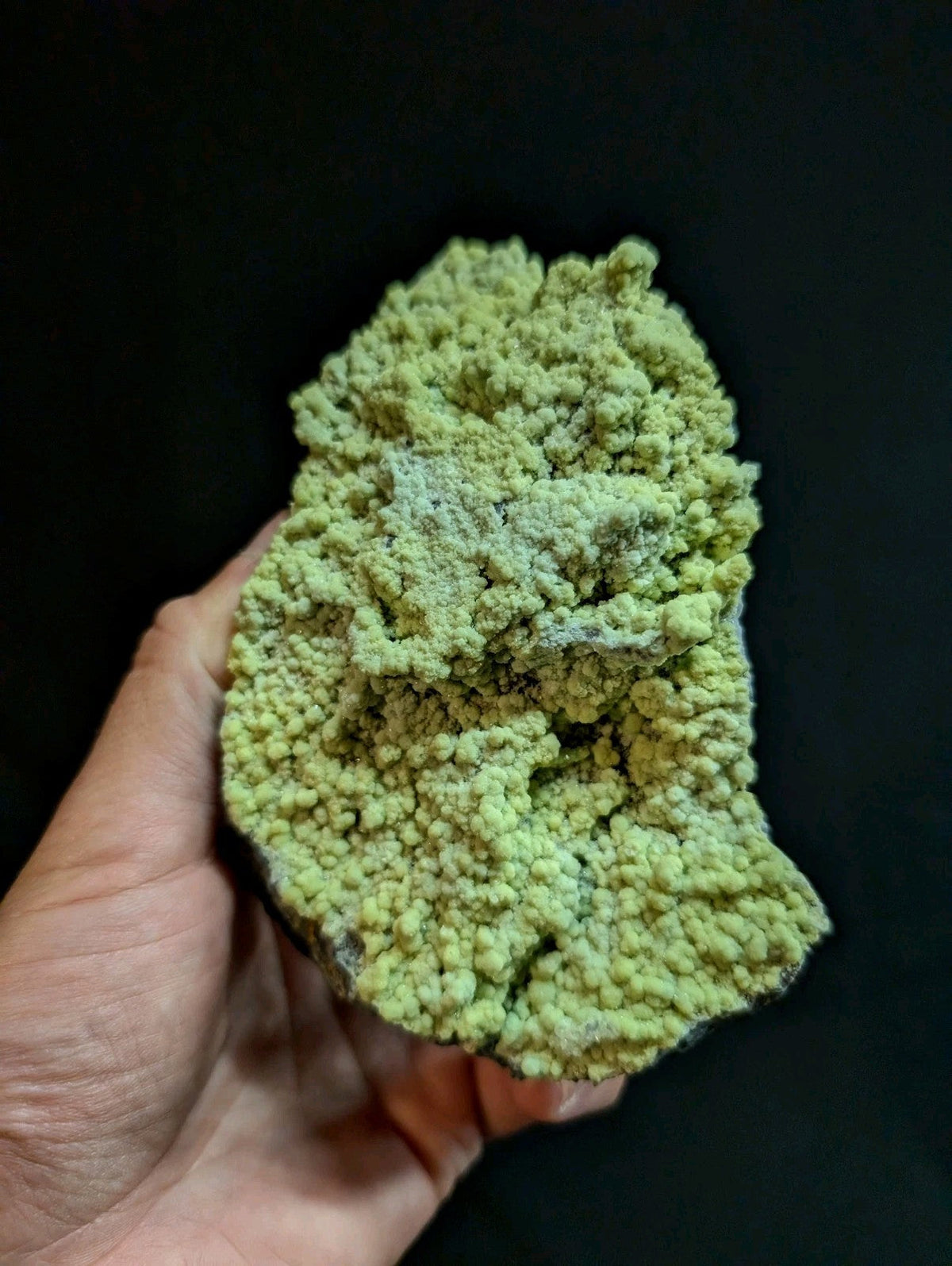 A hand holds a rough, textured specimen of greenish-yellow copper carbonate mineral with a crystalline, bumpy surface.