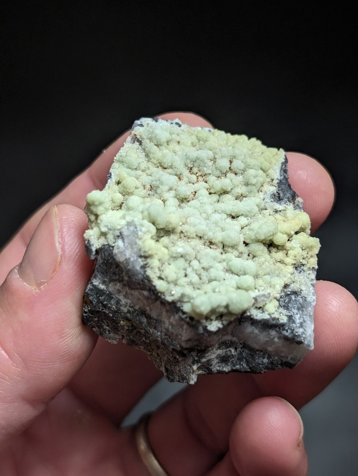 A hand holds a rough, irregularly shaped mineral specimen featuring a dense cluster of pale greenish-yellow nodules atop a darker gray rock base.