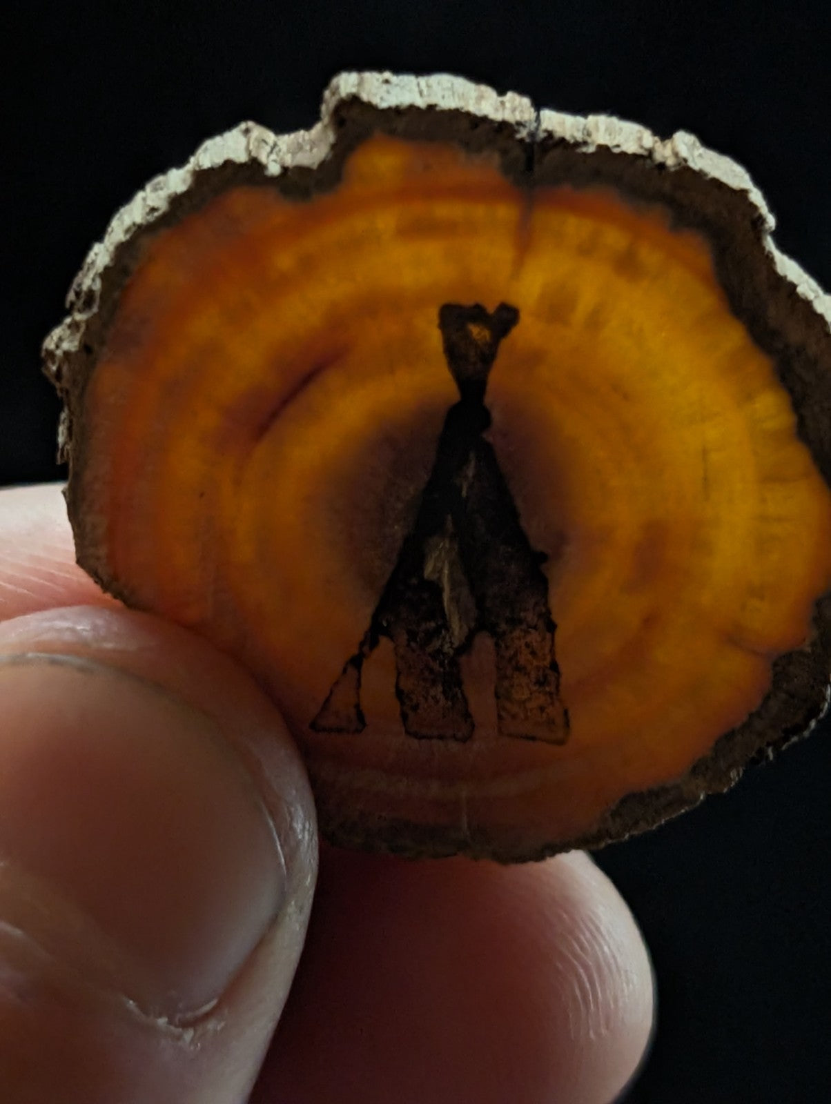 A circular, amber-hued wood slice pendant featuring a darkly engraved tribal-style figure design.