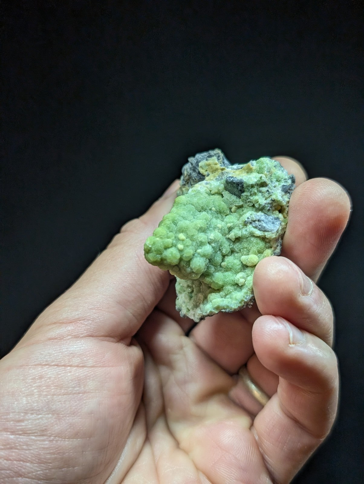 A hand holds a rough, irregularly shaped green mineral specimen with small nodules and patches of darker grayish material.