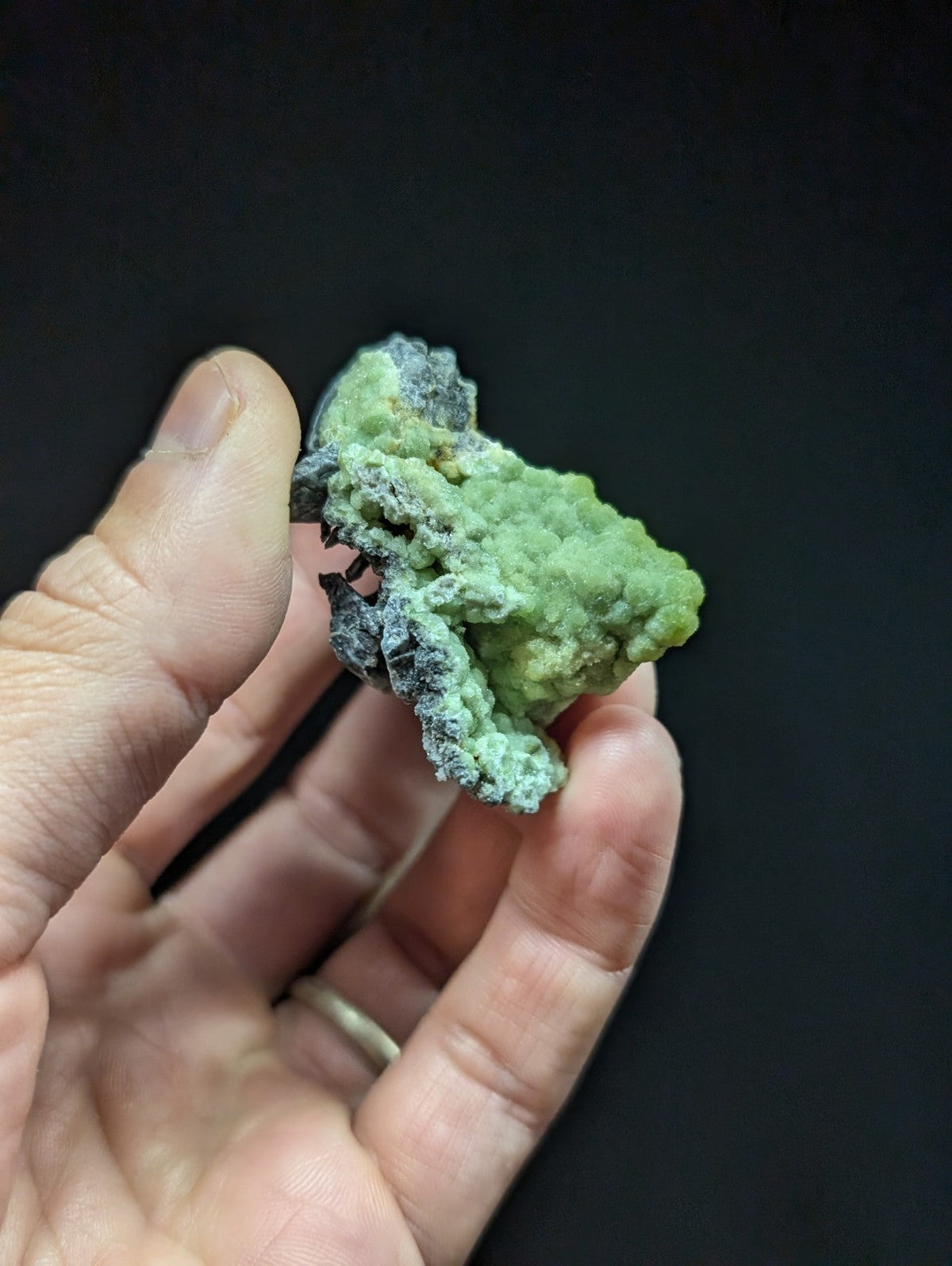 A hand holds a rough, irregularly shaped green mineral specimen with patches of dark gray and black veining.