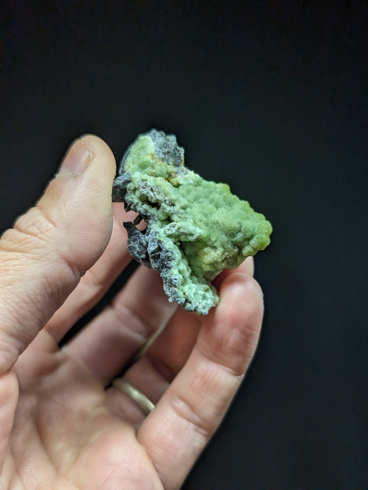 A hand holds a rough, irregularly shaped green mineral specimen with patches of dark gray and black veining.
