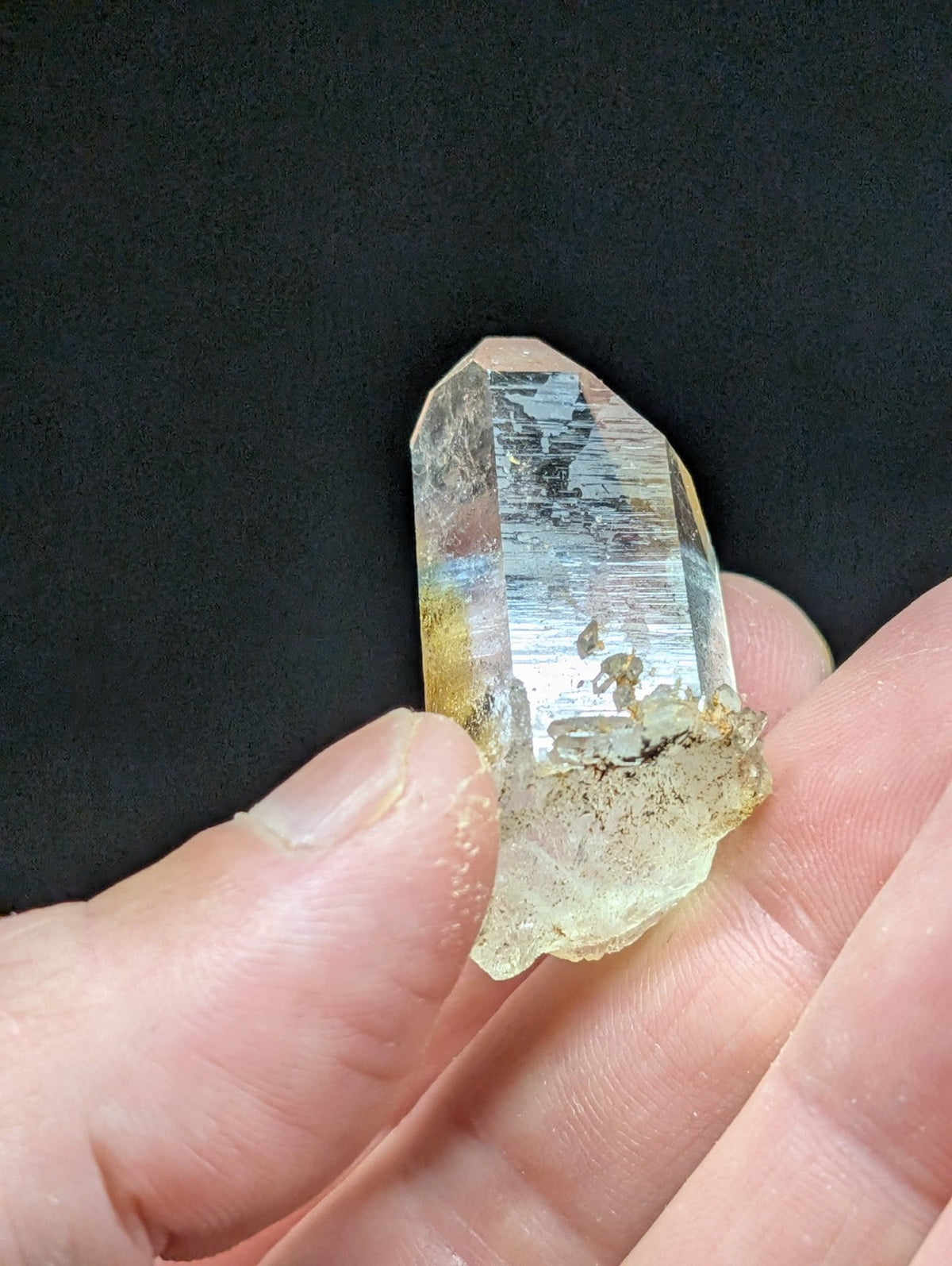 A translucent, clear quartz crystal with a pointed tip and natural inclusions, held gently between fingers against a dark background.