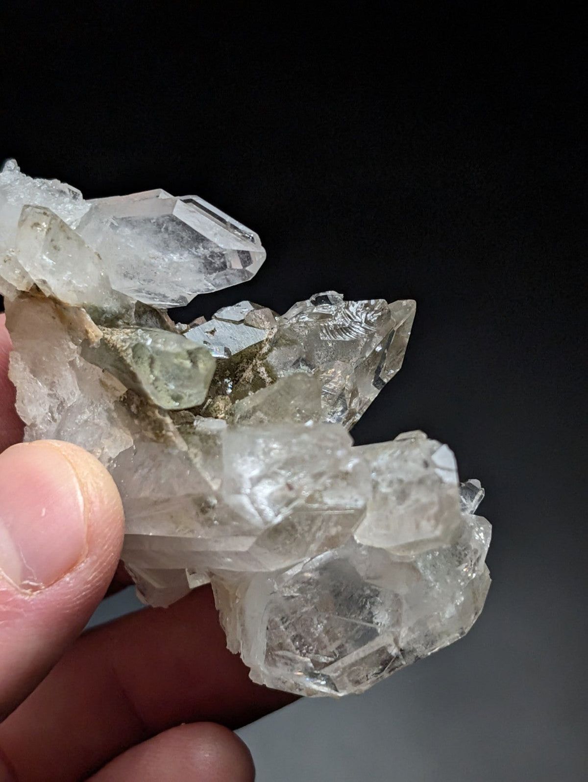 A cluster of clear, faceted quartz crystals with some translucent greenish hues and rougher surrounding rock.