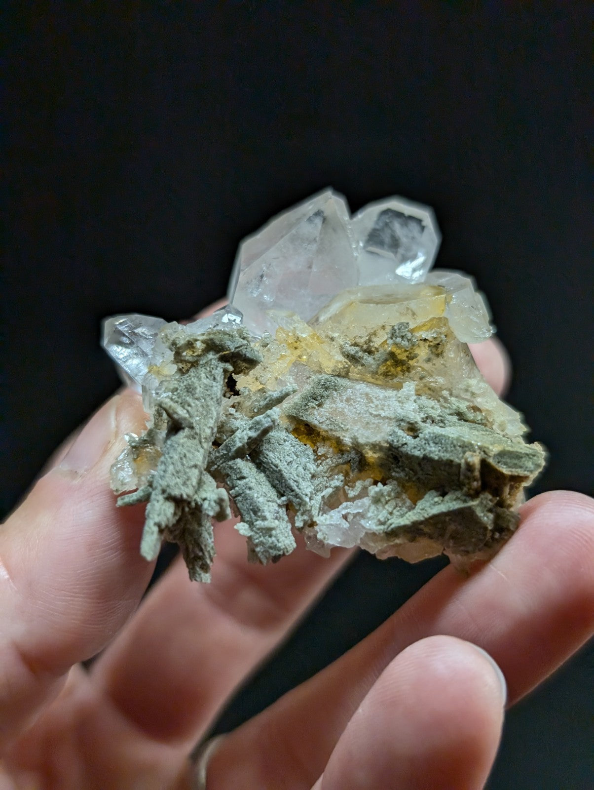 A hand holds a geological specimen featuring translucent quartz crystals with greenish-gray mineral matrix and yellow inclusions.