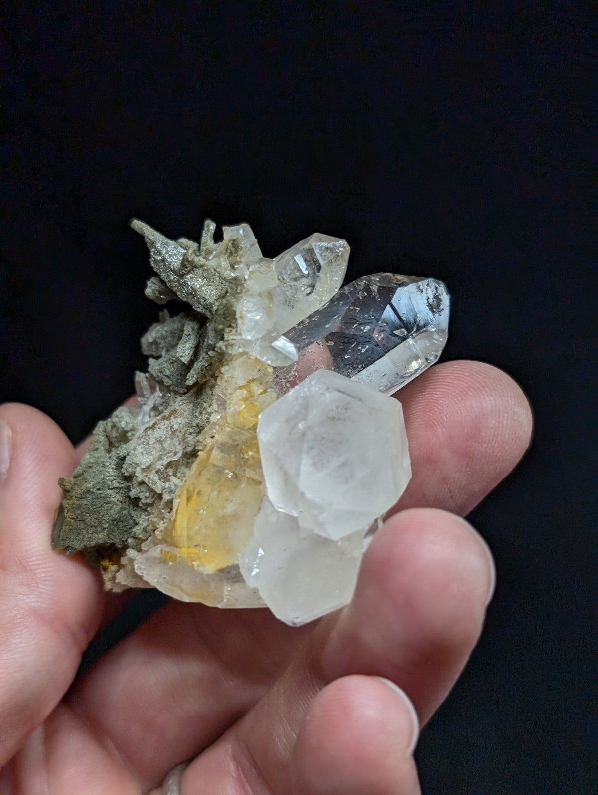 A hand holds a cluster of clear quartz crystals with some yellowish and translucent formations, set against a dark background.