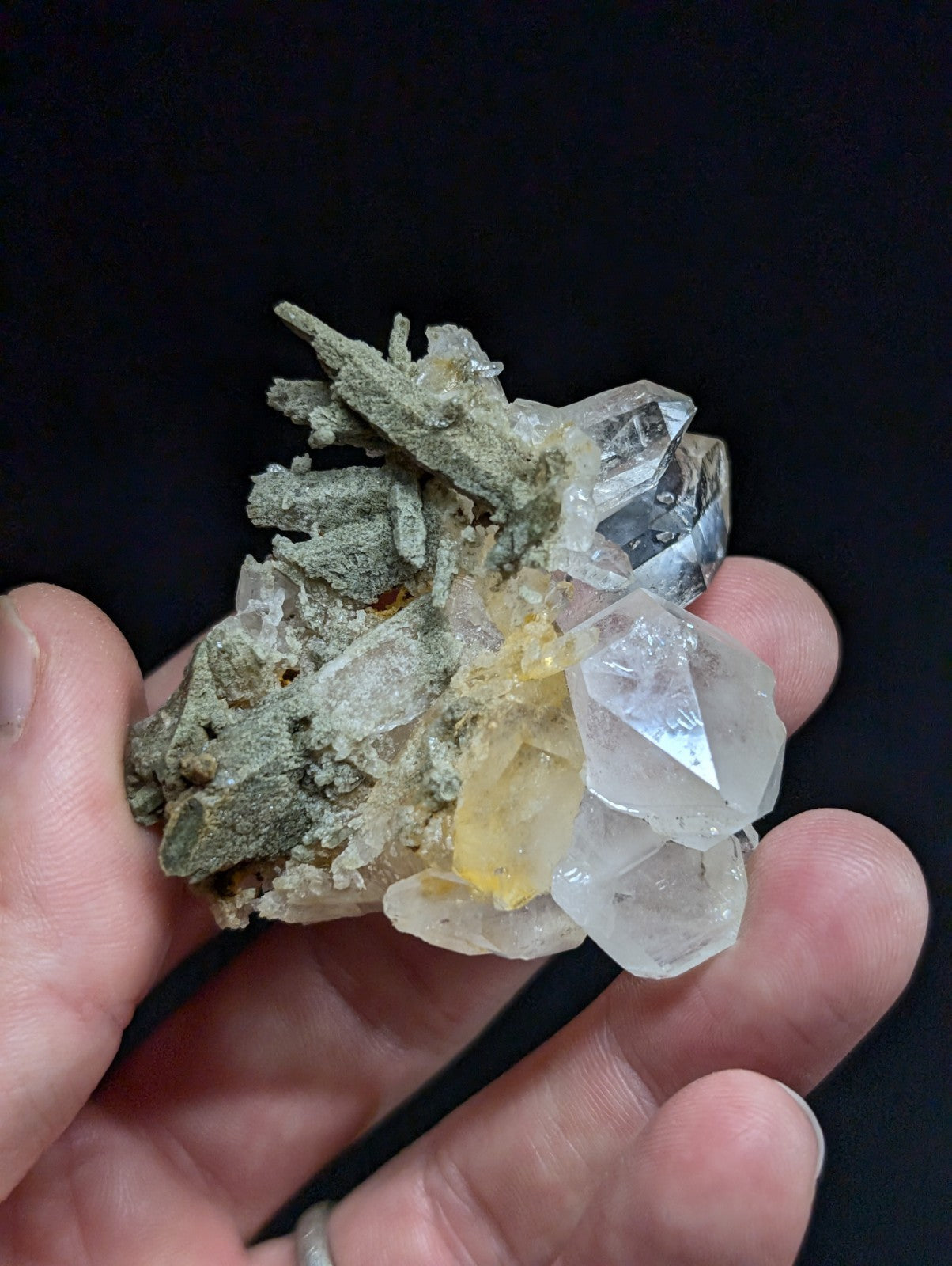 A hand holds a mineral specimen featuring clear, faceted quartz crystals with one yellowish inclusion and greenish-gray matrix material.