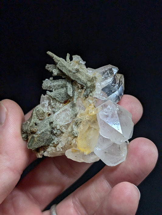A hand holds a mineral specimen featuring clear, faceted quartz crystals with one yellowish inclusion and greenish-gray matrix material.