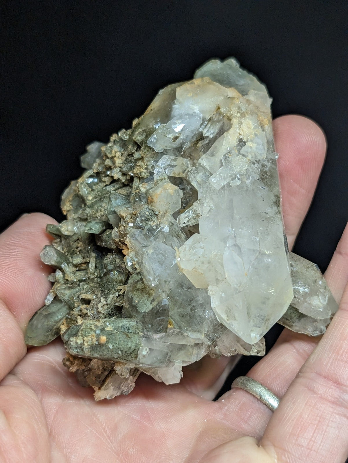 A hand holds a cluster of translucent quartz crystals with clear, faceted points and earthy greenish-gray matrix material.