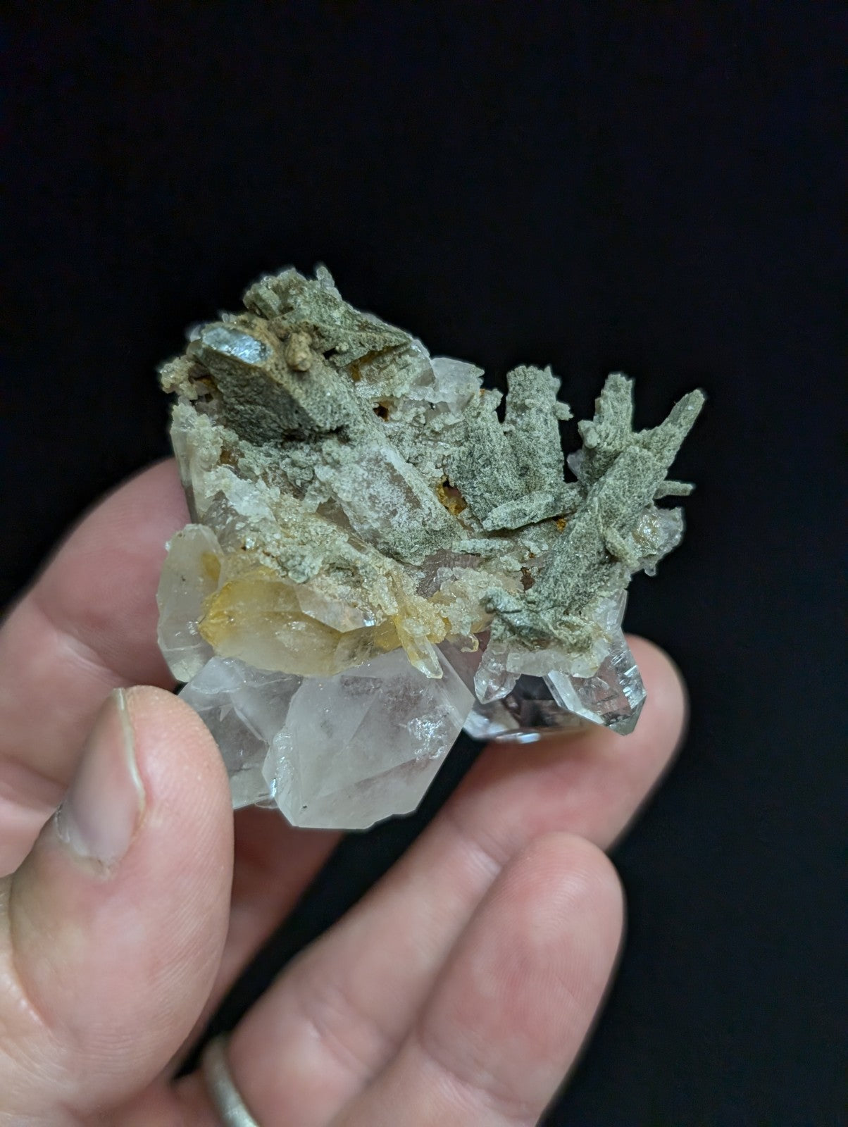 A hand holds a mineral specimen featuring translucent white quartz crystals with greenish-gray metallic veining and a small yellow inclusion.