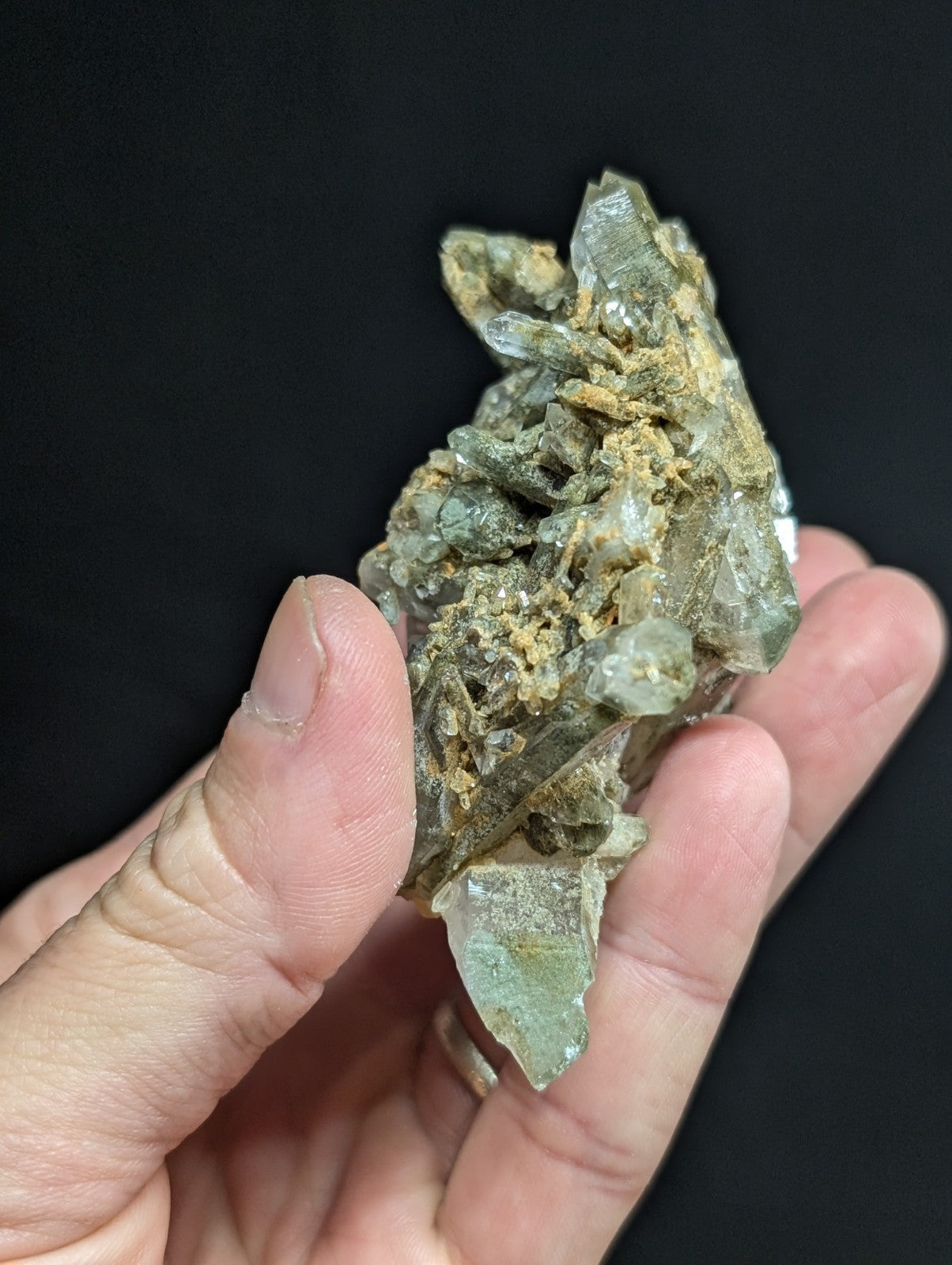 A hand holds a cluster of translucent greenish-gray quartz crystals with jagged, pointed facets and earthy brown matrix material.