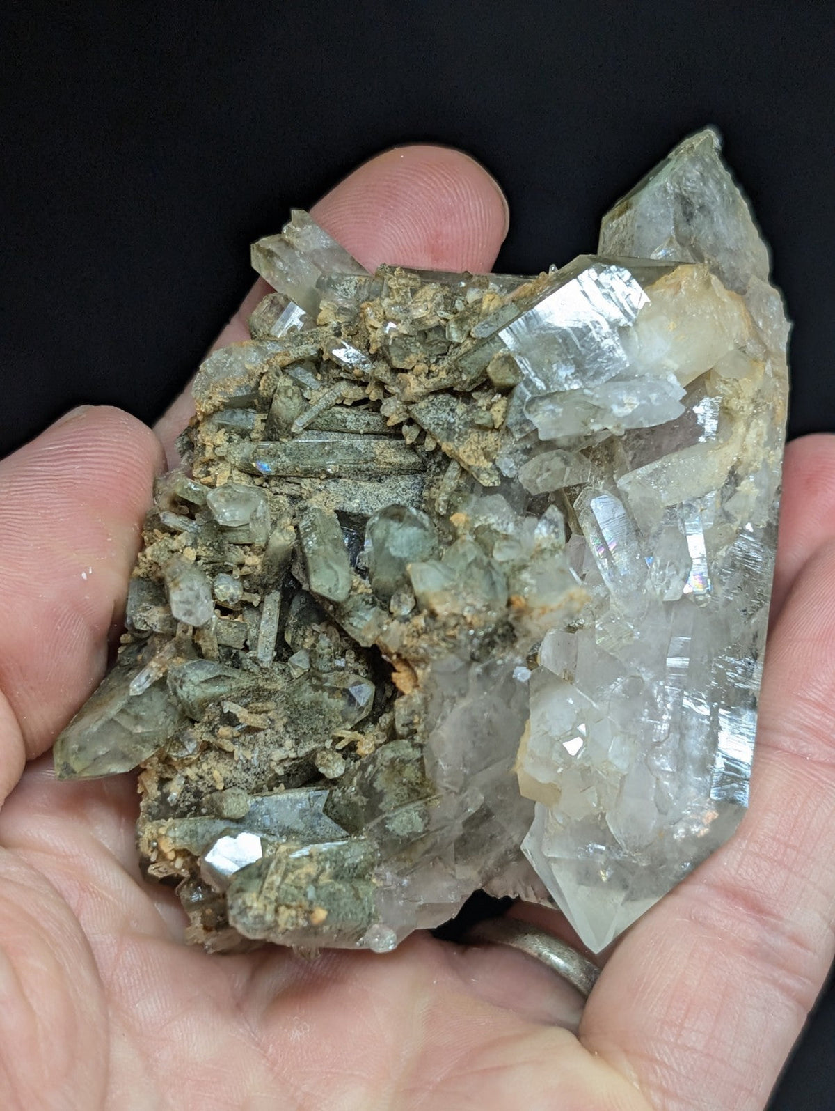 A hand holds a cluster of translucent green and clear quartz crystals with some exhibiting subtle iridescence, set against a dark background.