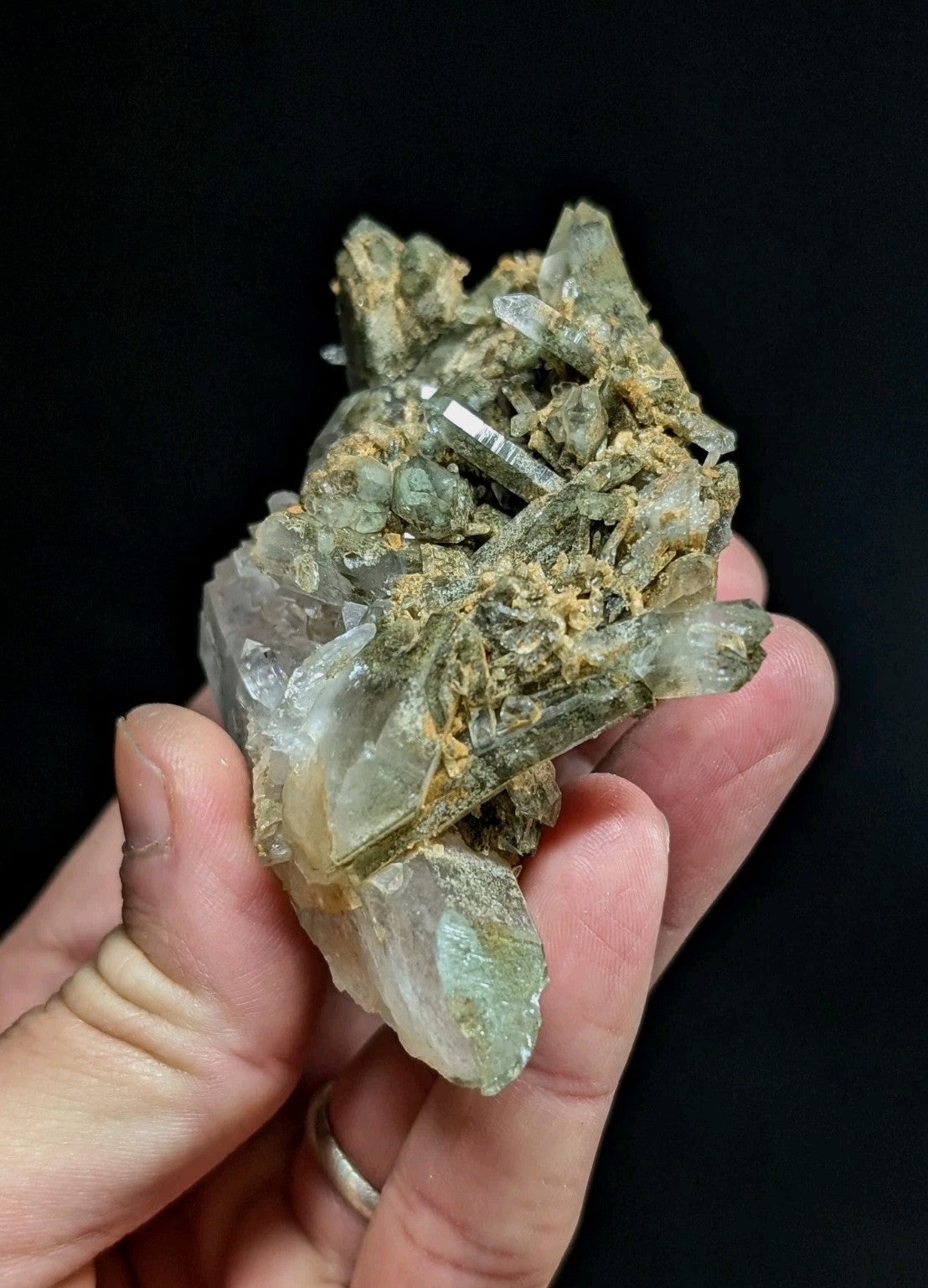 A hand holds a cluster of translucent greenish quartz crystals with embedded metallic inclusions and earthy matrix.